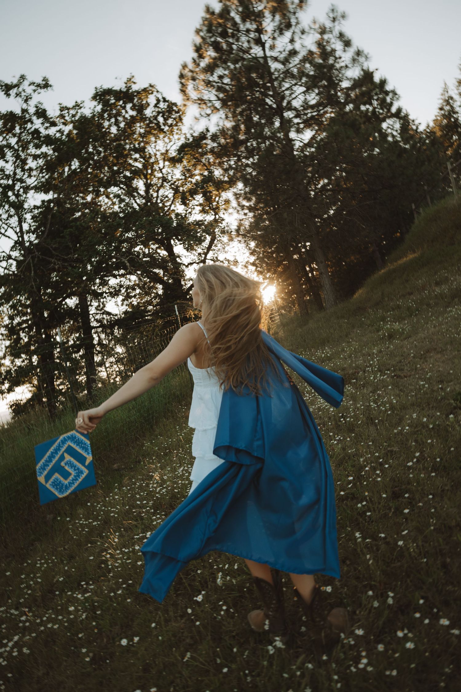 Blonde woman twirling in white dress with blue graduation gown in sunlit wildflower meadow surrounded by tall trees.