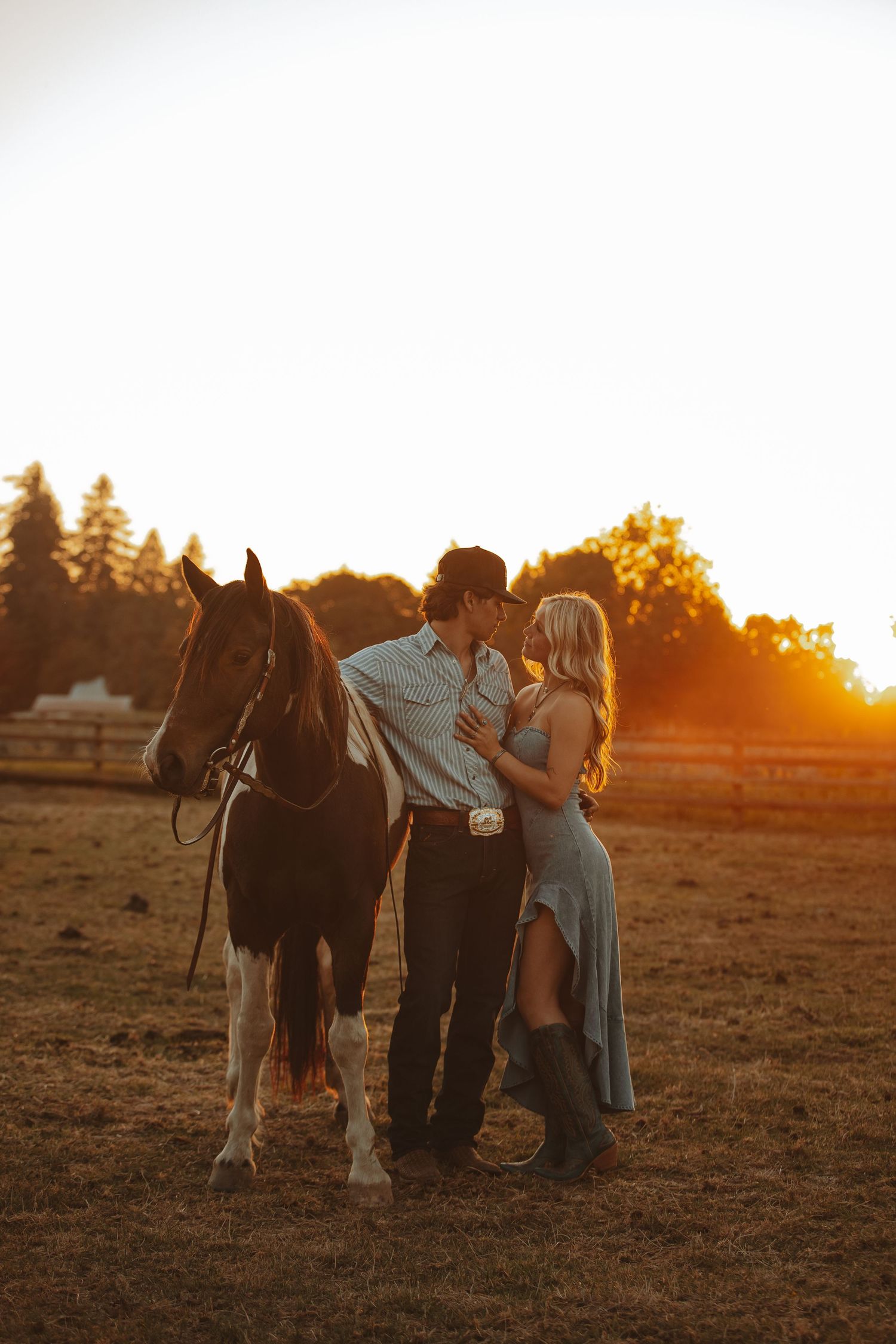 Couple sharing a romantic moment near a horse at golden hour sunset on a rustic farm field.