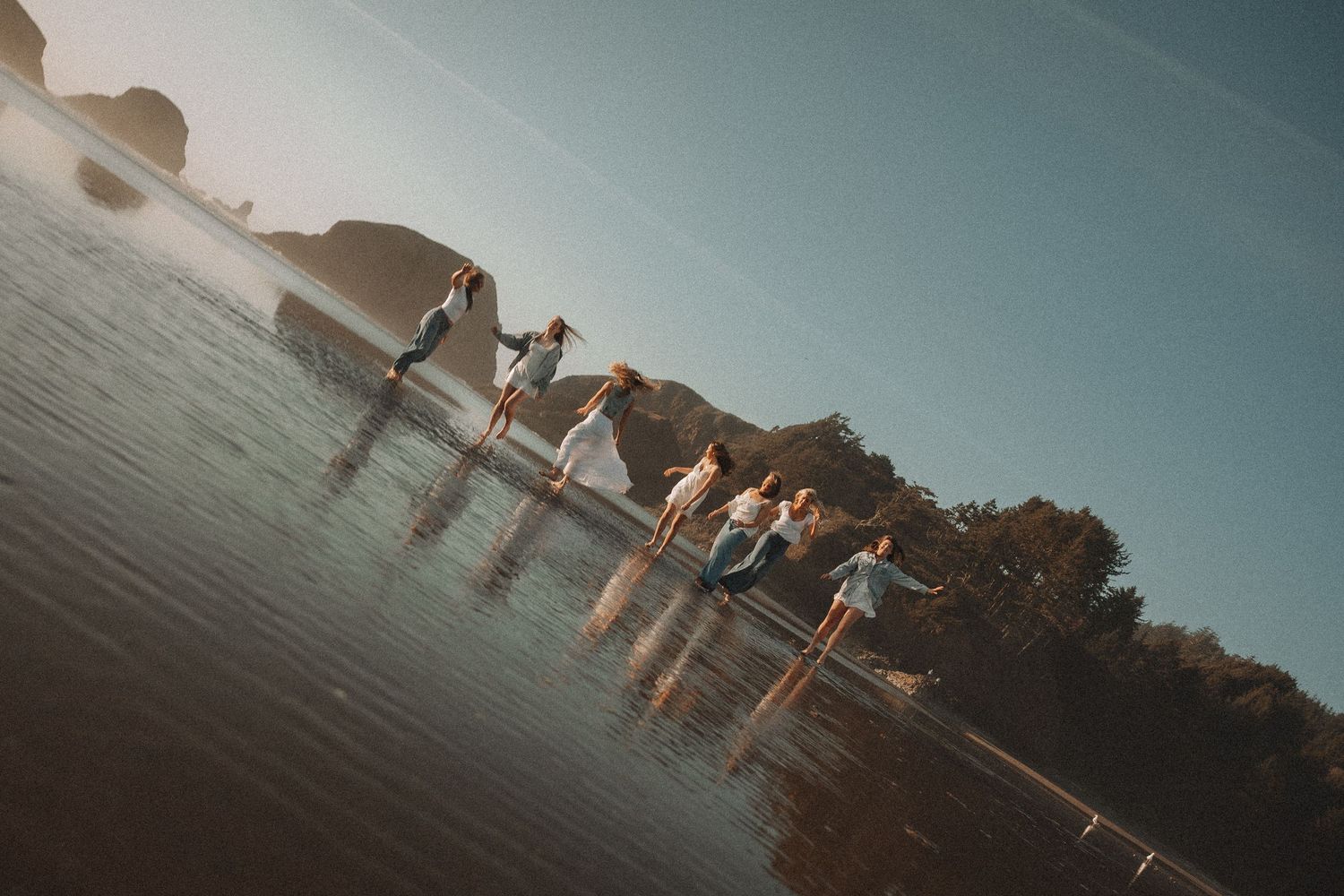 Group of friends holding hands walking in shallow water near a rocky shoreline at golden hour sunset.