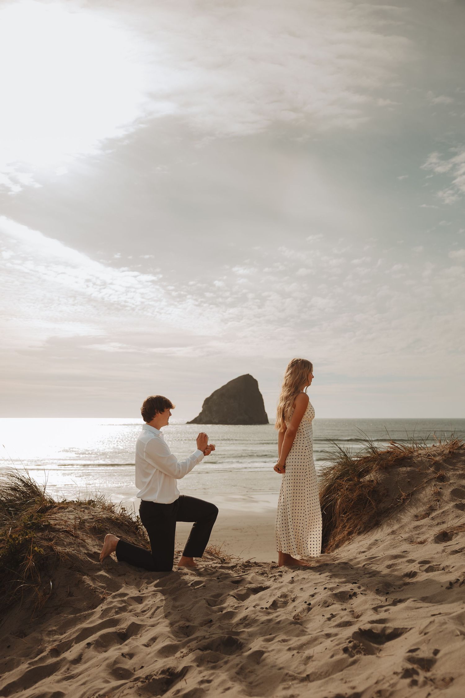 Man proposes to woman on sandy beach dunes at sunset with rocky outcrop in background.