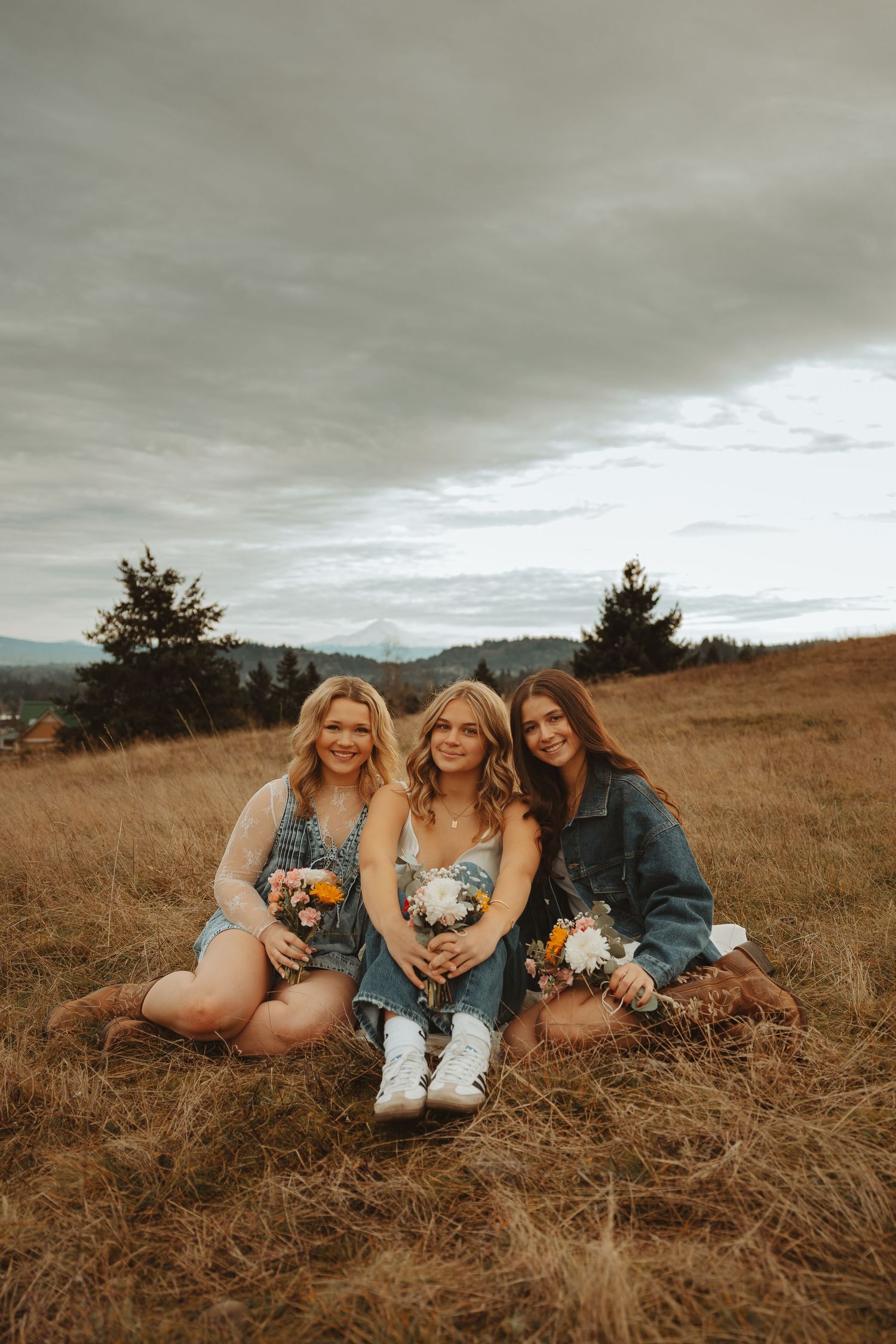 Three friends relax together in a rustic field wearing casual denim and white dresses.