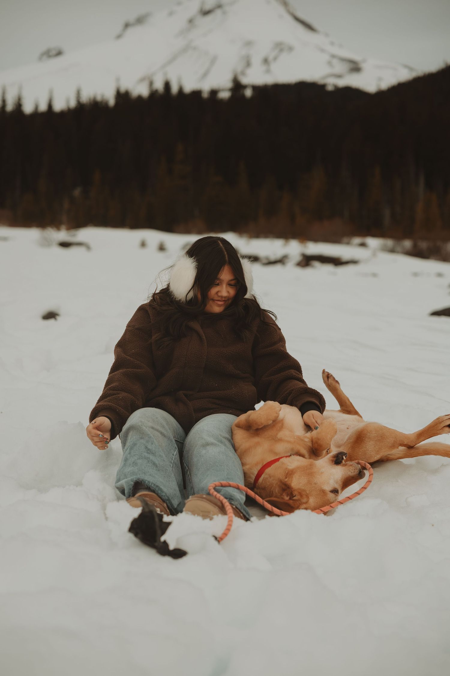 Woman in brown hoodie sitting in snow playing with a golden dog near a snowy mountain.