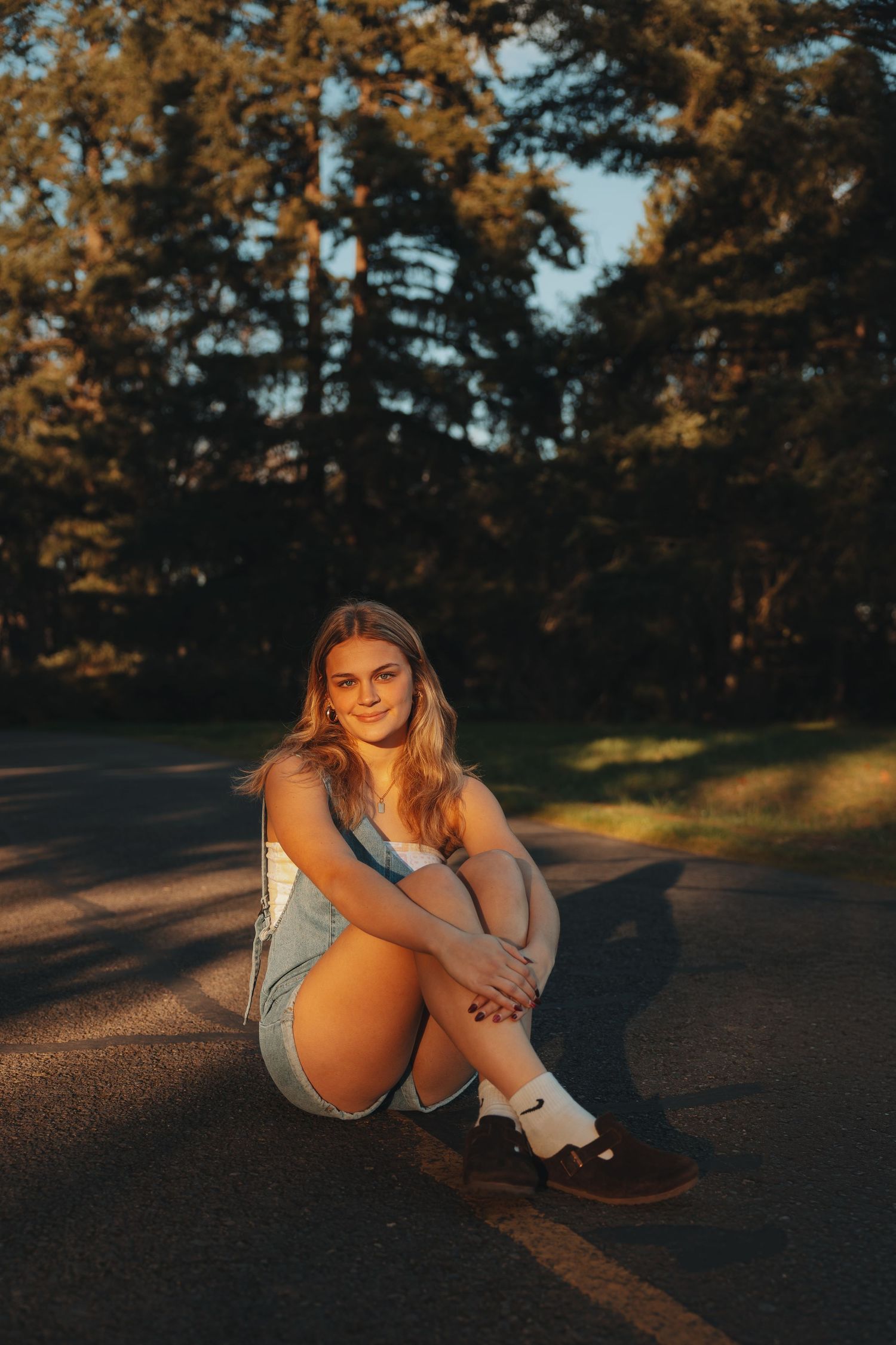 Young woman sitting on a skateboard on a road at golden hour, wearing denim overalls, smiling at camera.