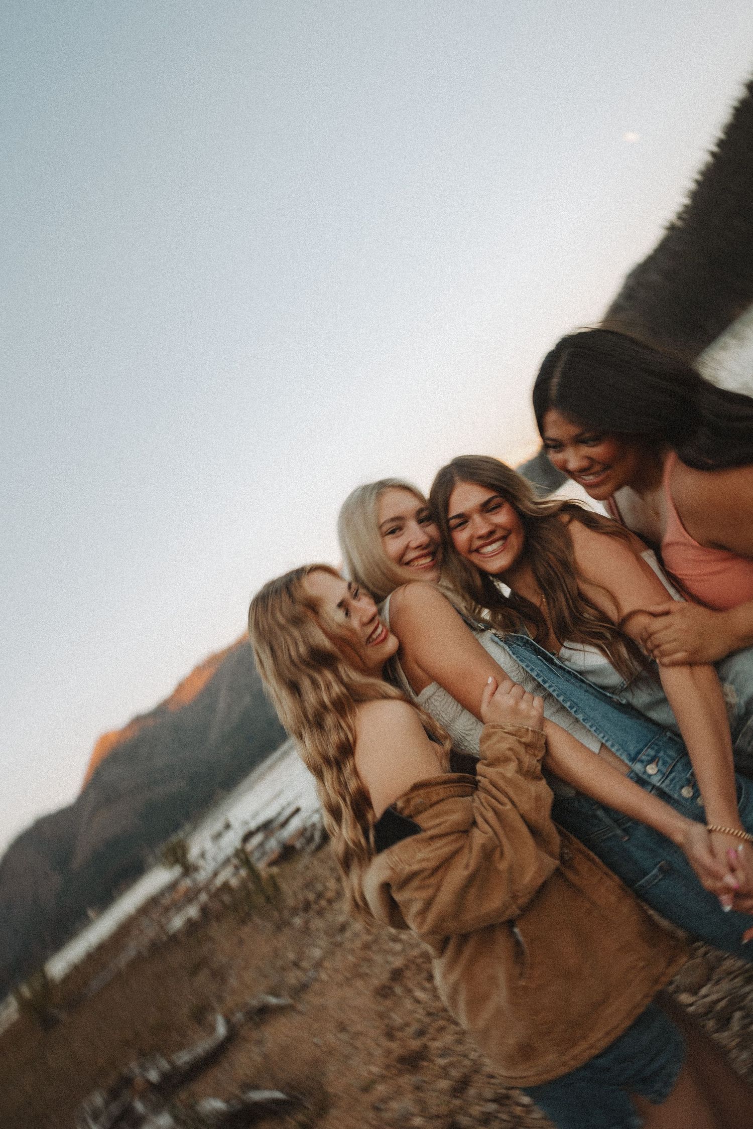 Four young women laughing and embracing on a rocky beach with mountain lake backdrop.