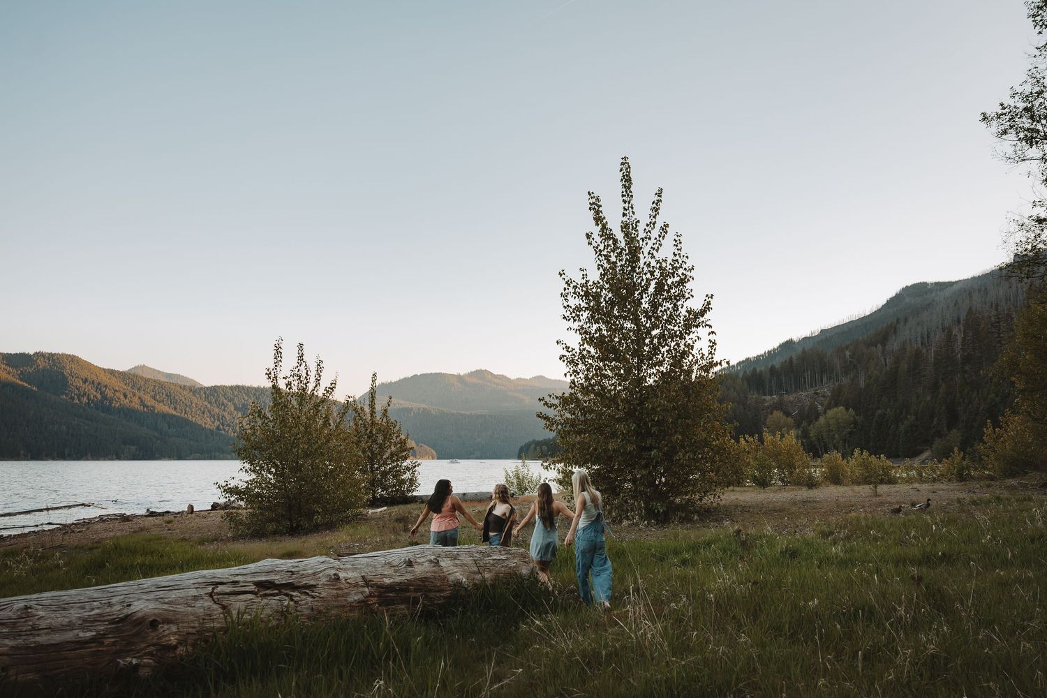 Group of young women gathered near a fallen log by a mountain lake at dusk.