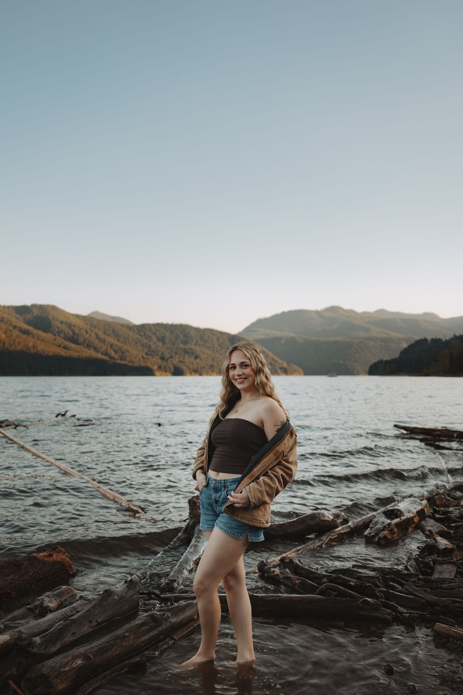 Smiling young woman in black top and denim shorts standing by a mountain lake at sunset.