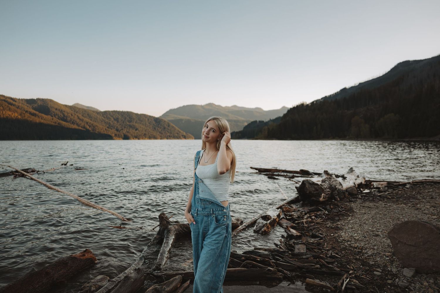 Woman in blue jeans standing by a serene mountain lake shore with driftwood at golden hour.