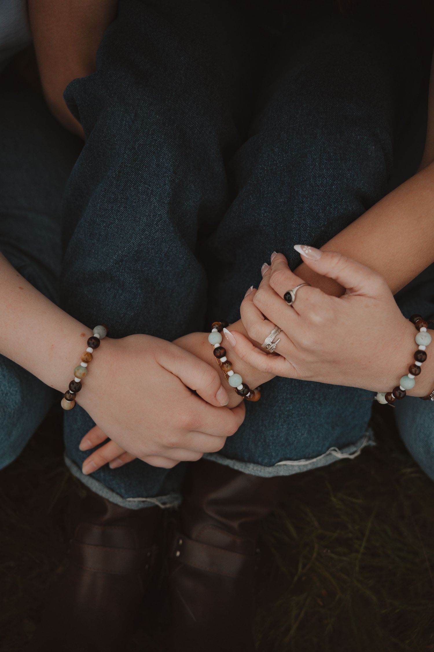 Close-up of two hands wearing beaded bracelets and rings, resting on denim jeans with brown boots.
