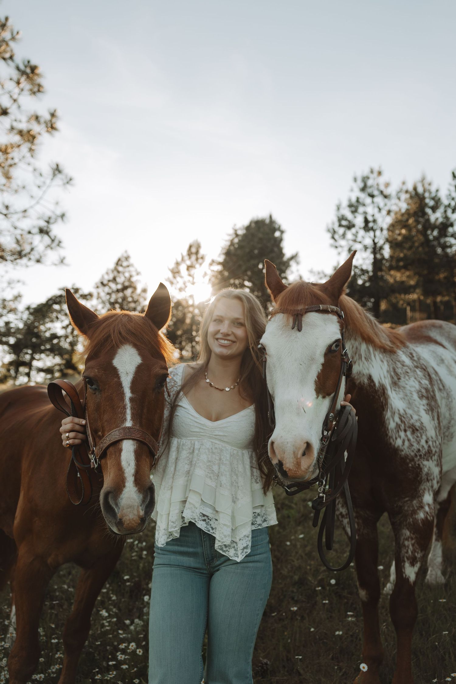 Smiling young woman standing between two chestnut horses in a sunlit forest field, wearing a white lace top and jeans.
