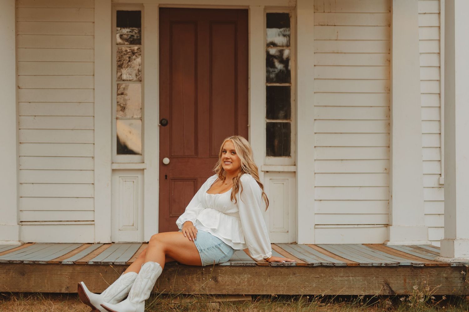 Young blonde woman in white top, denim skirt, and white boots sitting on a rustic porch smiling.