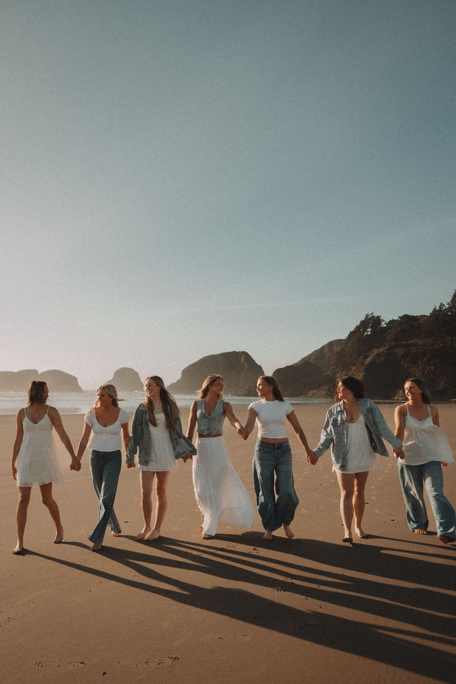 Seven young women in white and denim holding hands walking on a sunny beach.