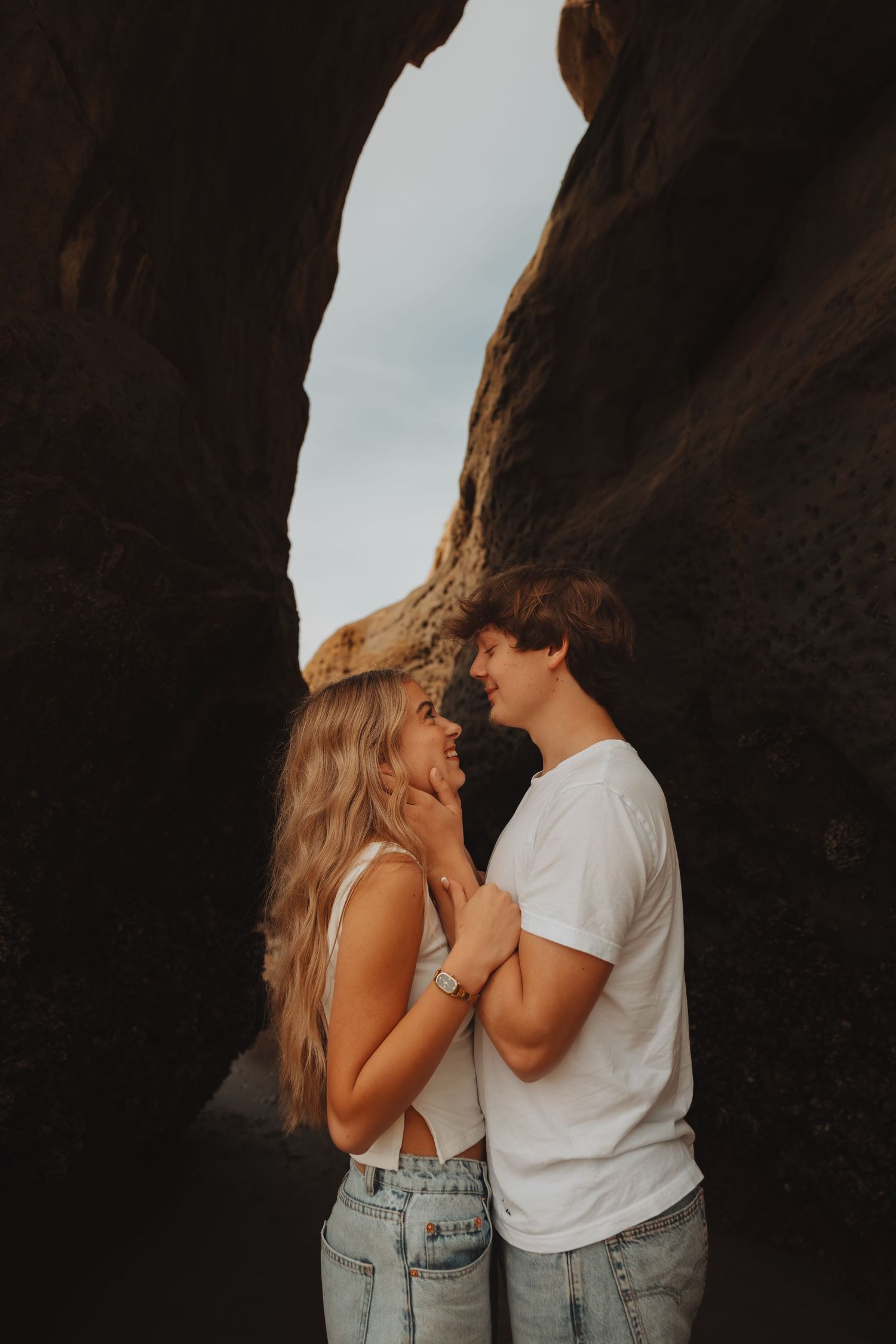 Romantic couple almost kissing while standing between towering dark rock formations on a beach.
