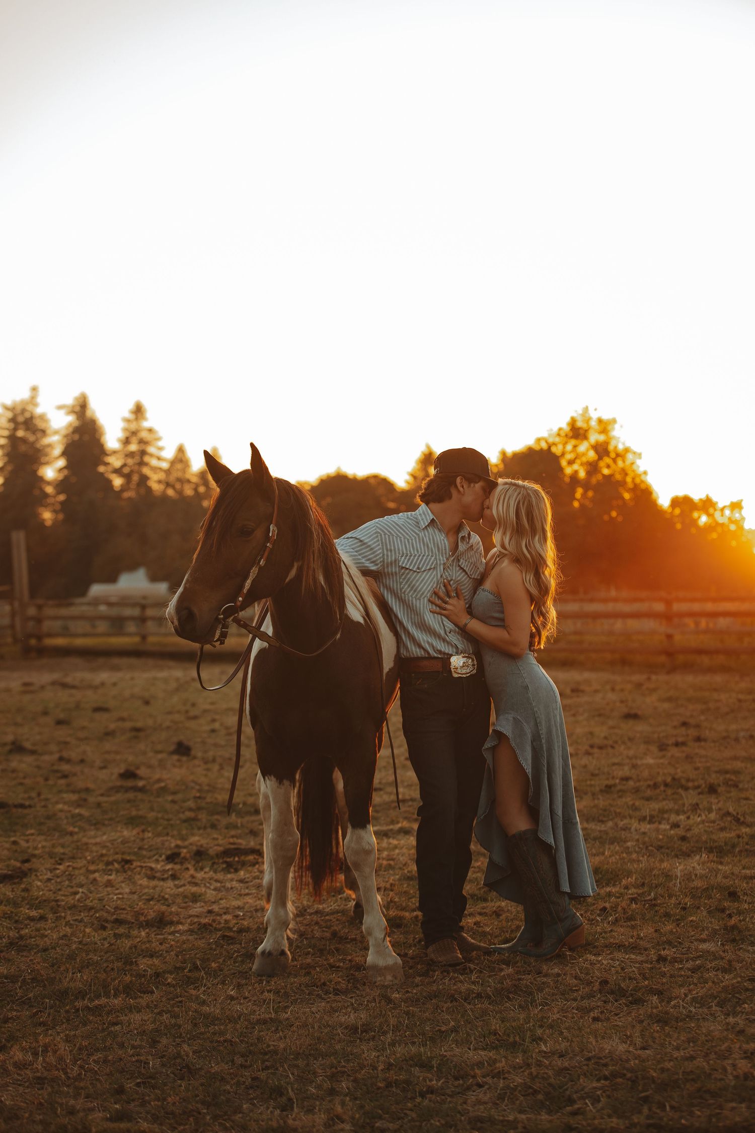 A couple poses with their horse in a field at golden sunset while wearing western attire.
