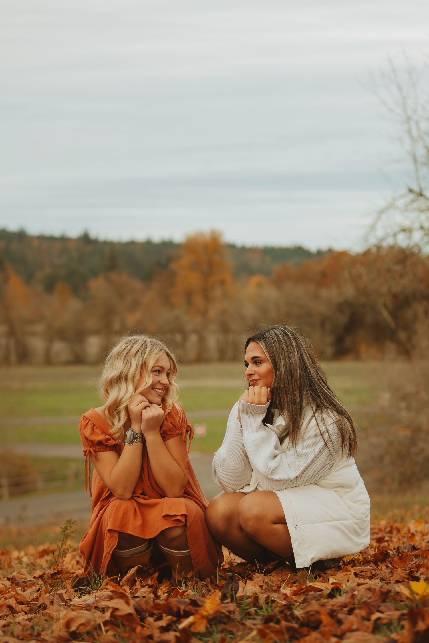 Two people lying in autumn leaves wearing orange and white dresses during a golden hour photoshoot.