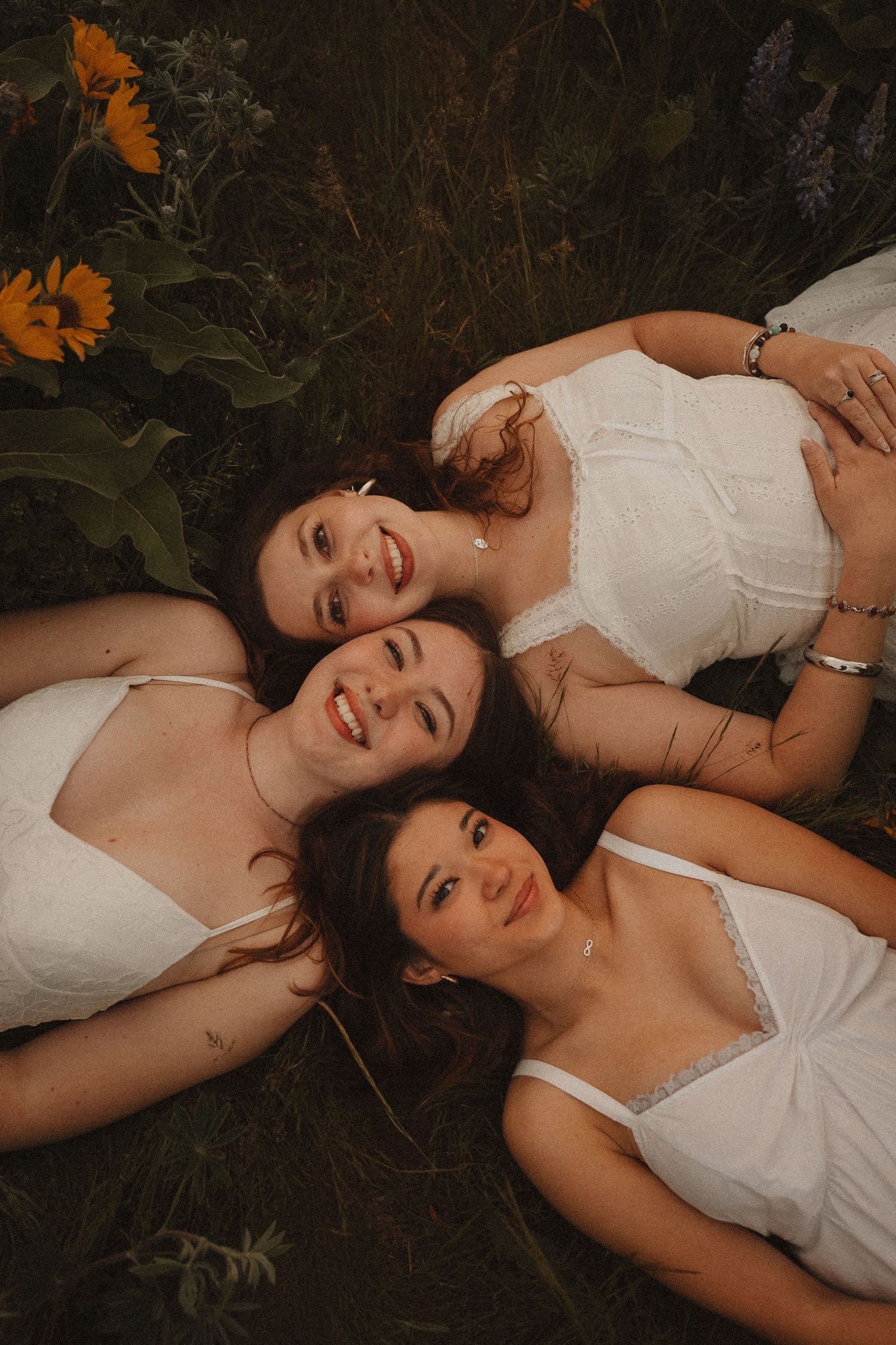 Three smiling women in white dresses lying together in a flower field with sunflowers and purple blooms.