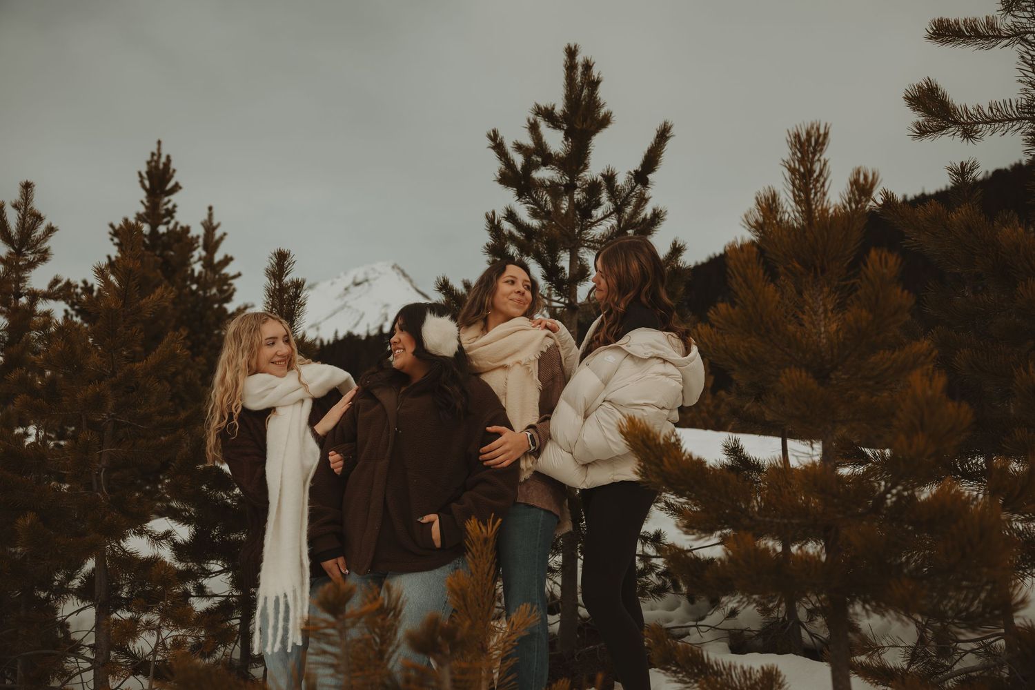 Four women laughing together among snow-dusted pine trees in winter mountain setting.
