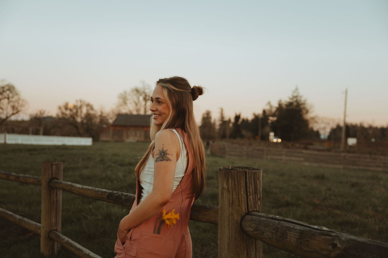Smiling young woman in pink overalls holding a flower leaning on a wooden fence at dusk.