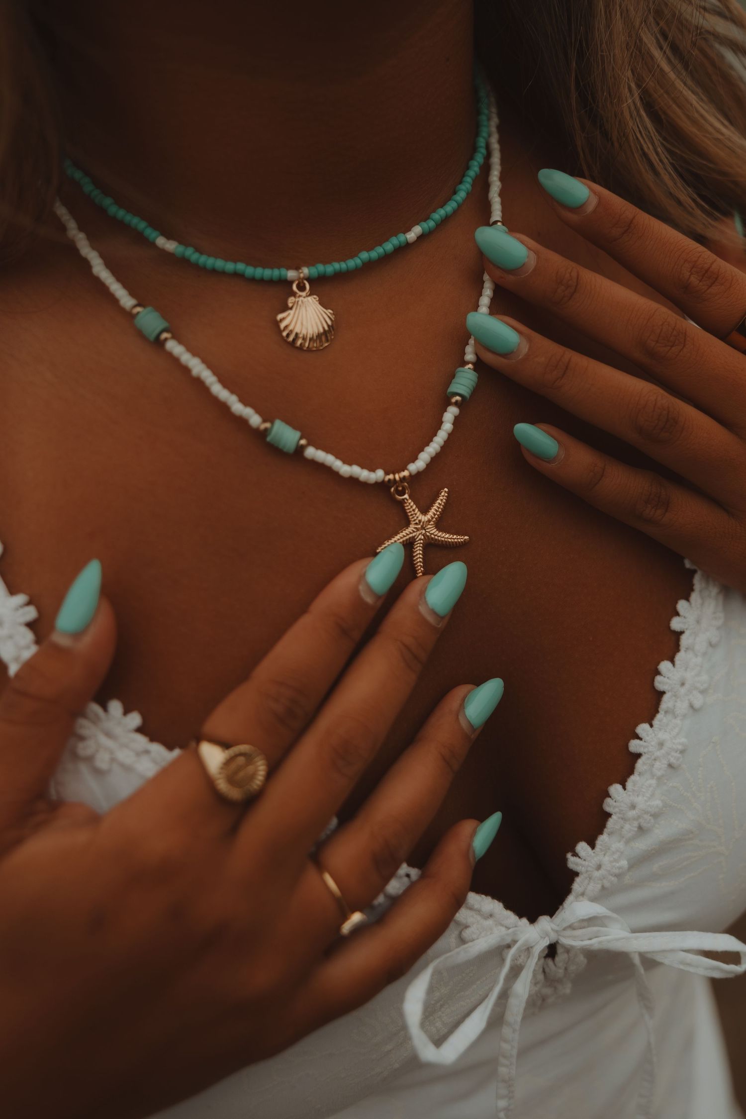 Woman wearing layered turquoise beaded necklaces with shell pendants, mint green nails, and gold rings.