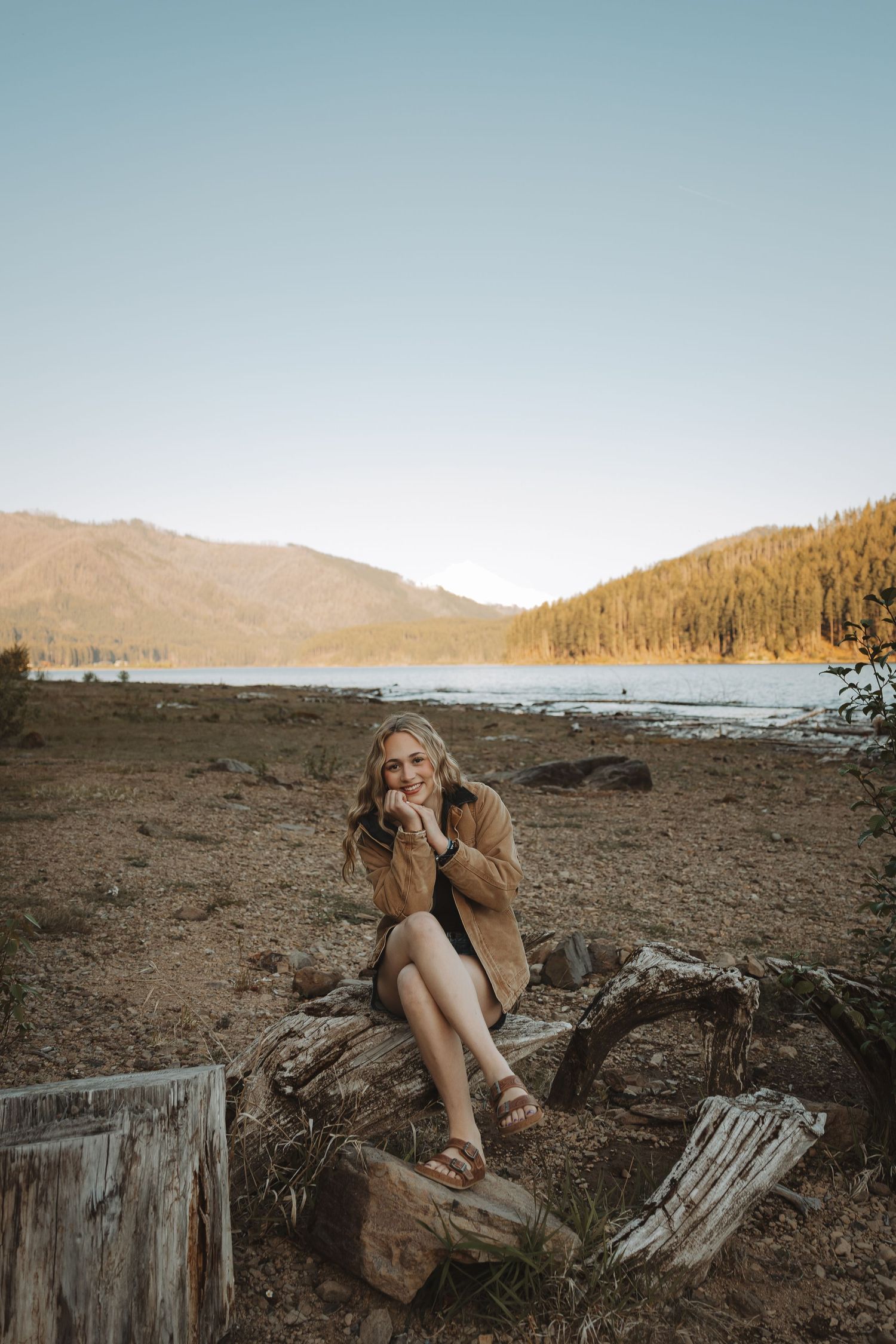 Young woman in brown jacket sits on driftwood logs by a mountain lake at golden hour.