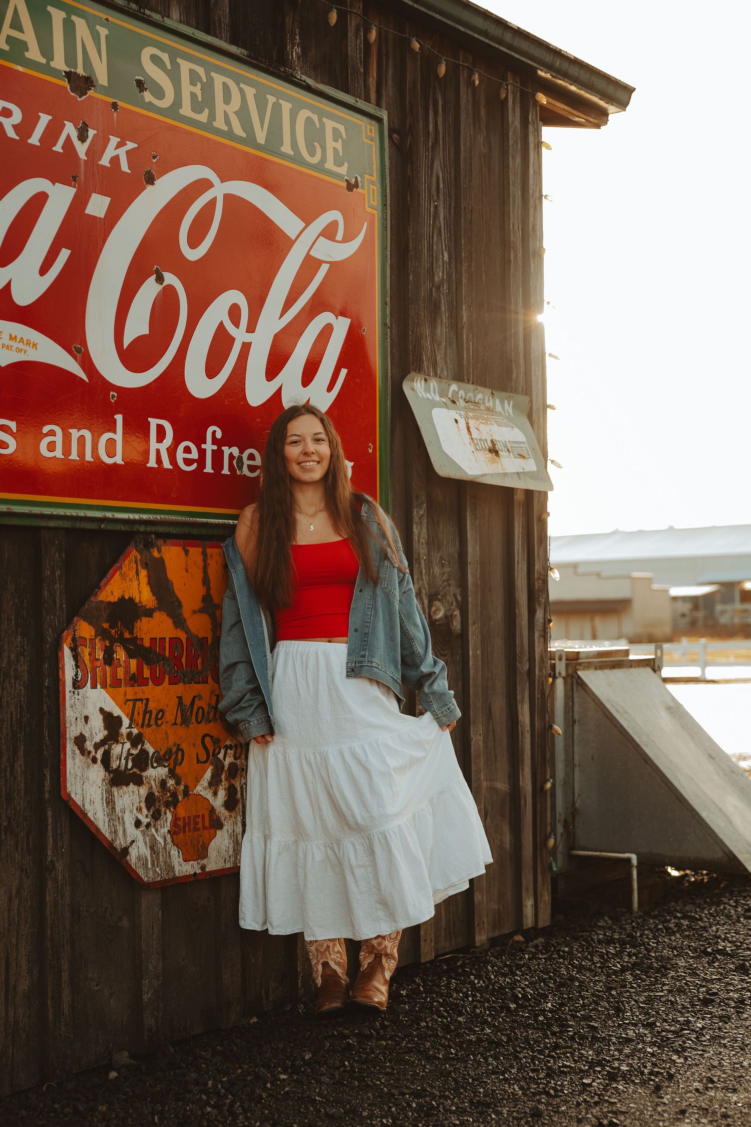 Smiling woman in red top, white midi skirt, and denim jacket leaning against rustic Coca-Cola sign in warm sunlight.
