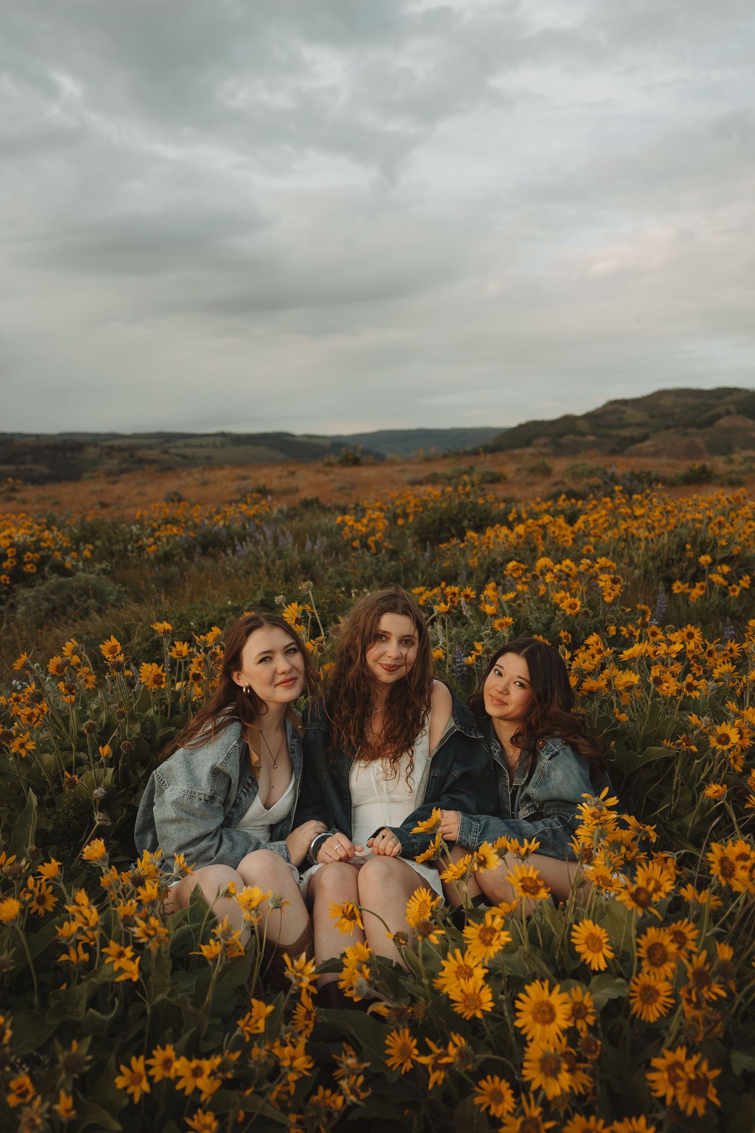 Three young women in denim jackets sit closely together in a golden wildflower meadow with rolling hills behind them.
