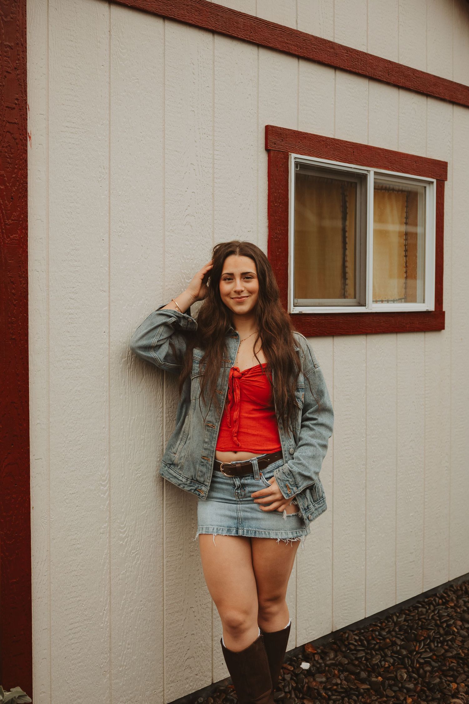 A person wearing a denim jacket and red top poses casually against a white building with brown trim.