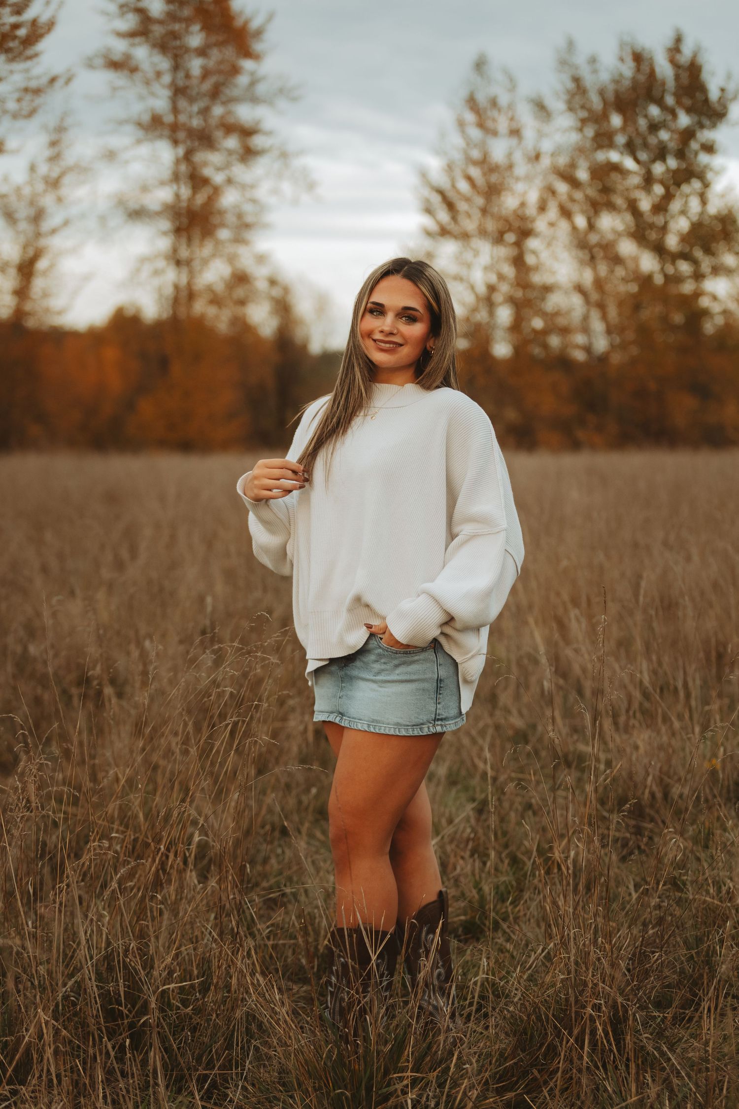 A woman in a white sweater and denim shorts poses in a golden autumn field during sunset.