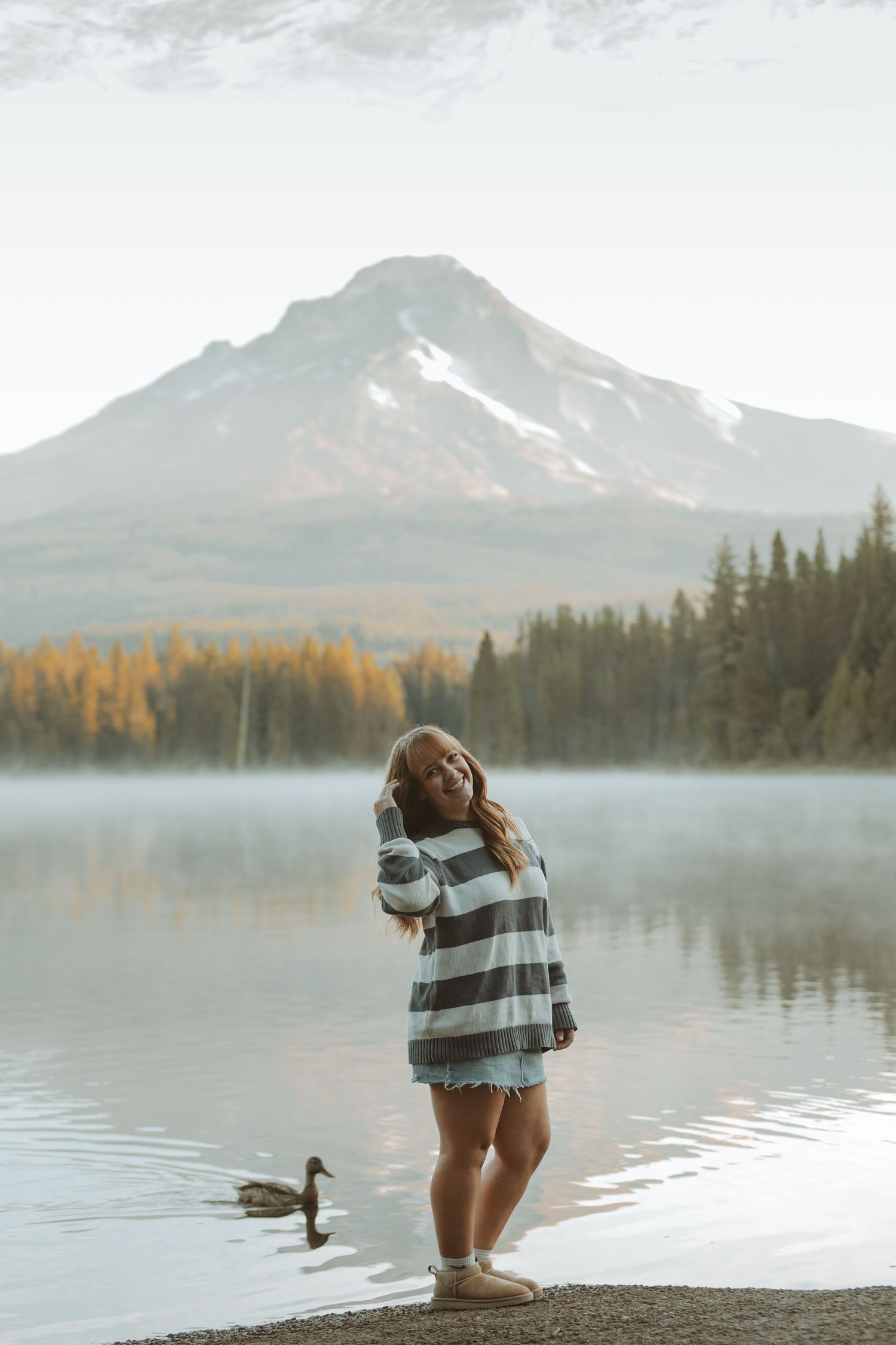 Misty morning at a mountain lake with pine trees and fall foliage reflecting in calm waters.