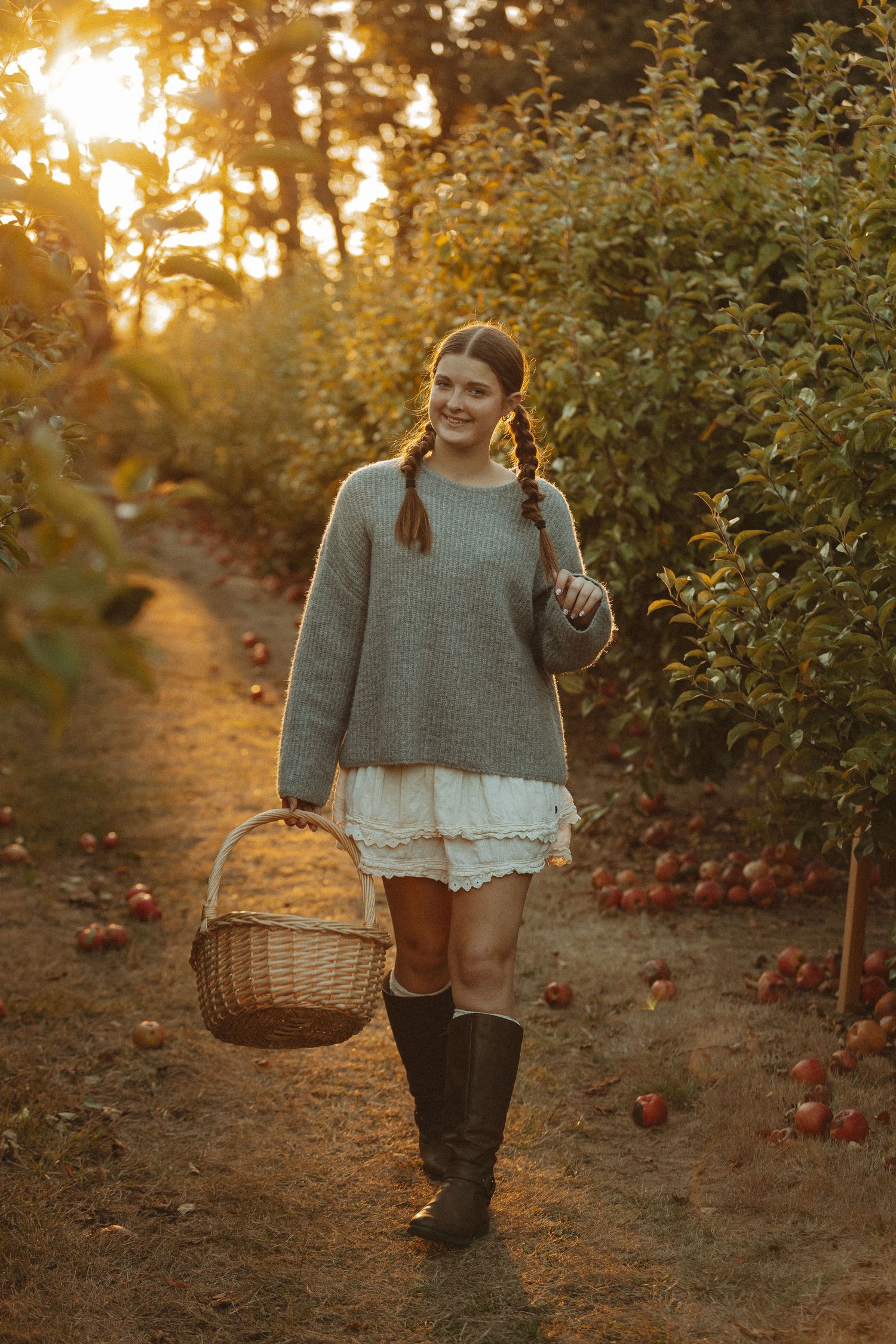A person in a gray sweater and boots carries a woven basket through an apple orchard at golden hour sunset.
