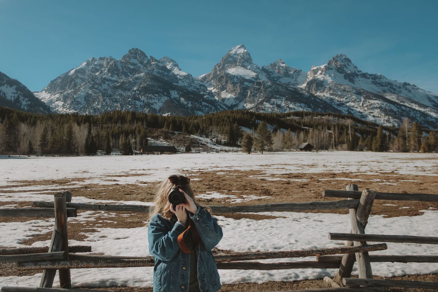 Woman photographing snowy Grand Teton mountain range behind rustic wooden fence on a clear winter day.