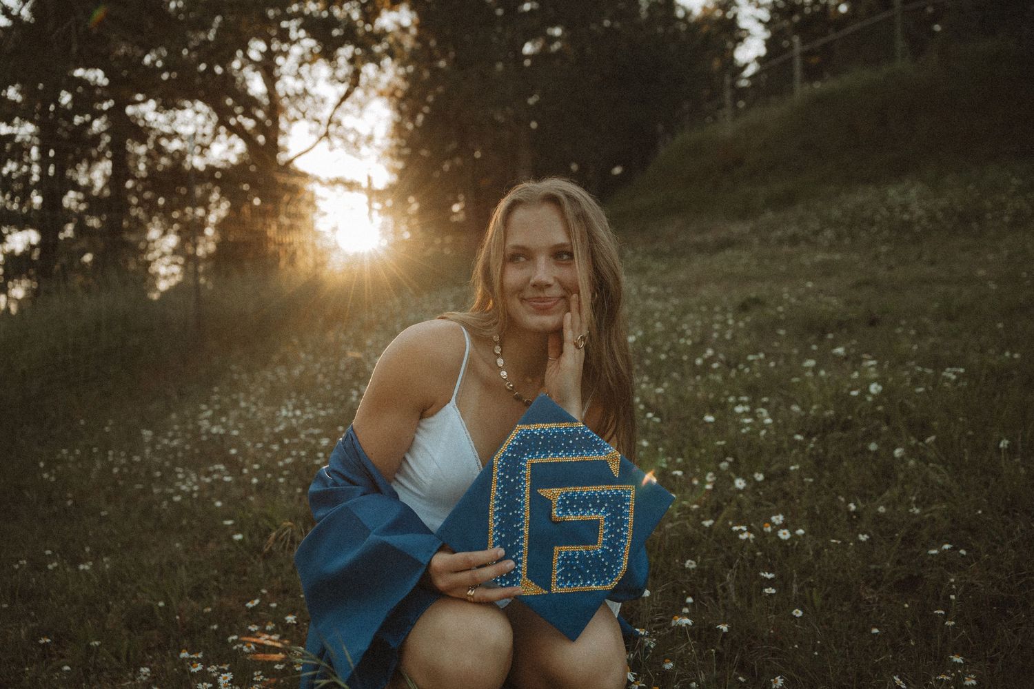 Woman holding blue decorative letter board at sunset in a flower field, wearing white top with blue wrap.
