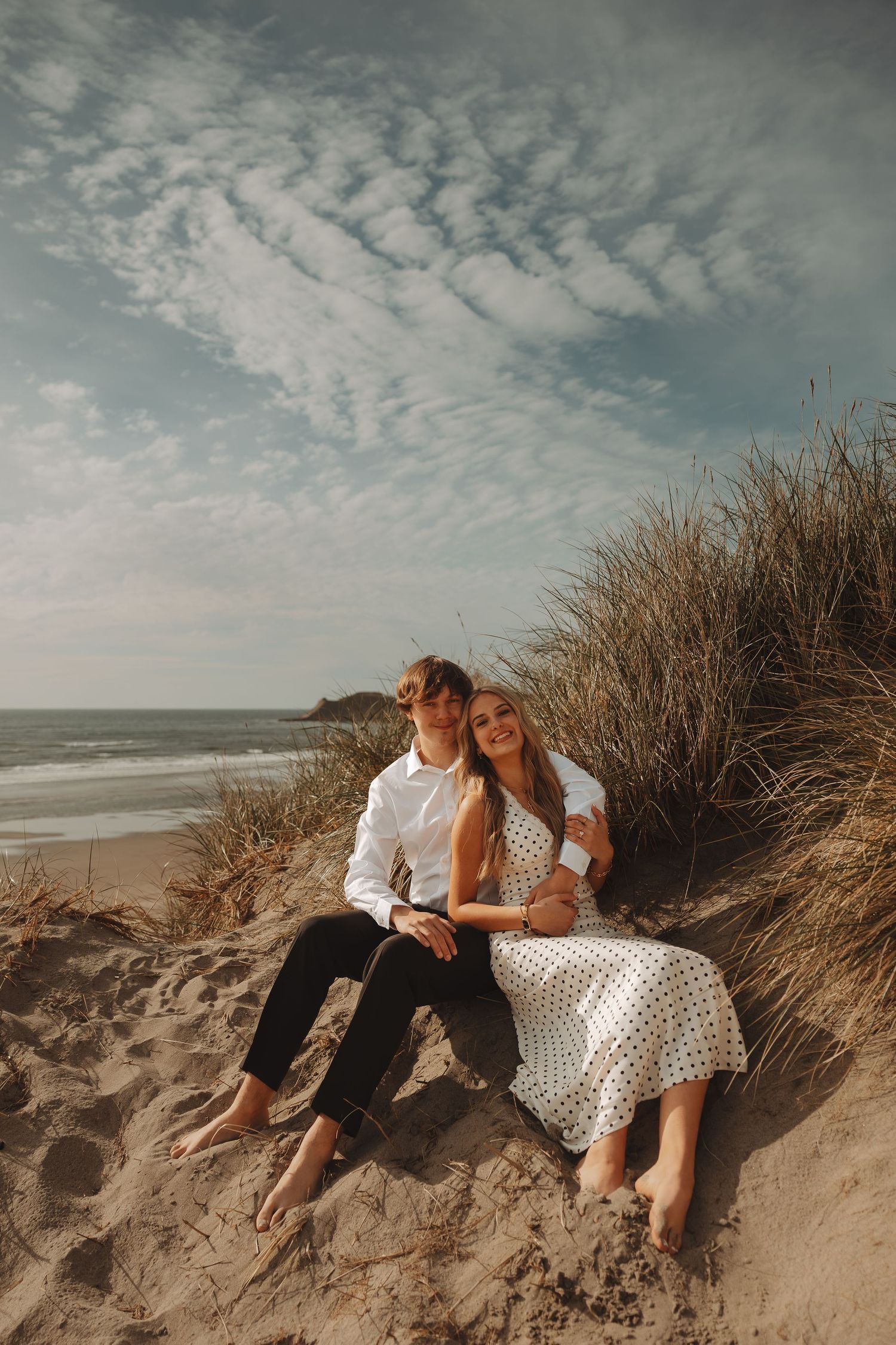 Happy couple sits together on sandy beach dunes surrounded by tall grass under a cloudy sky.