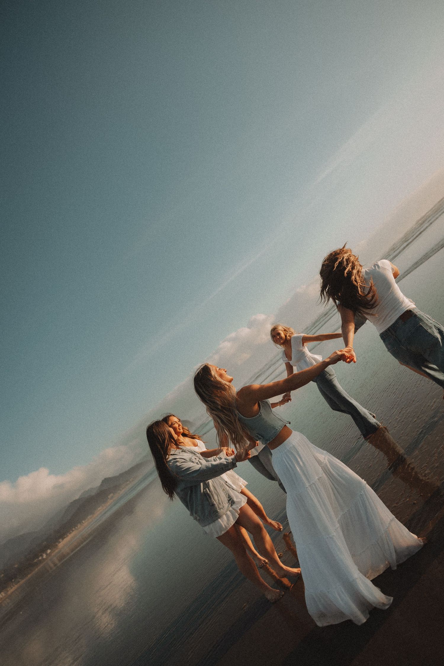 Three women in boho dresses joyfully dancing together on a beach at sunset.