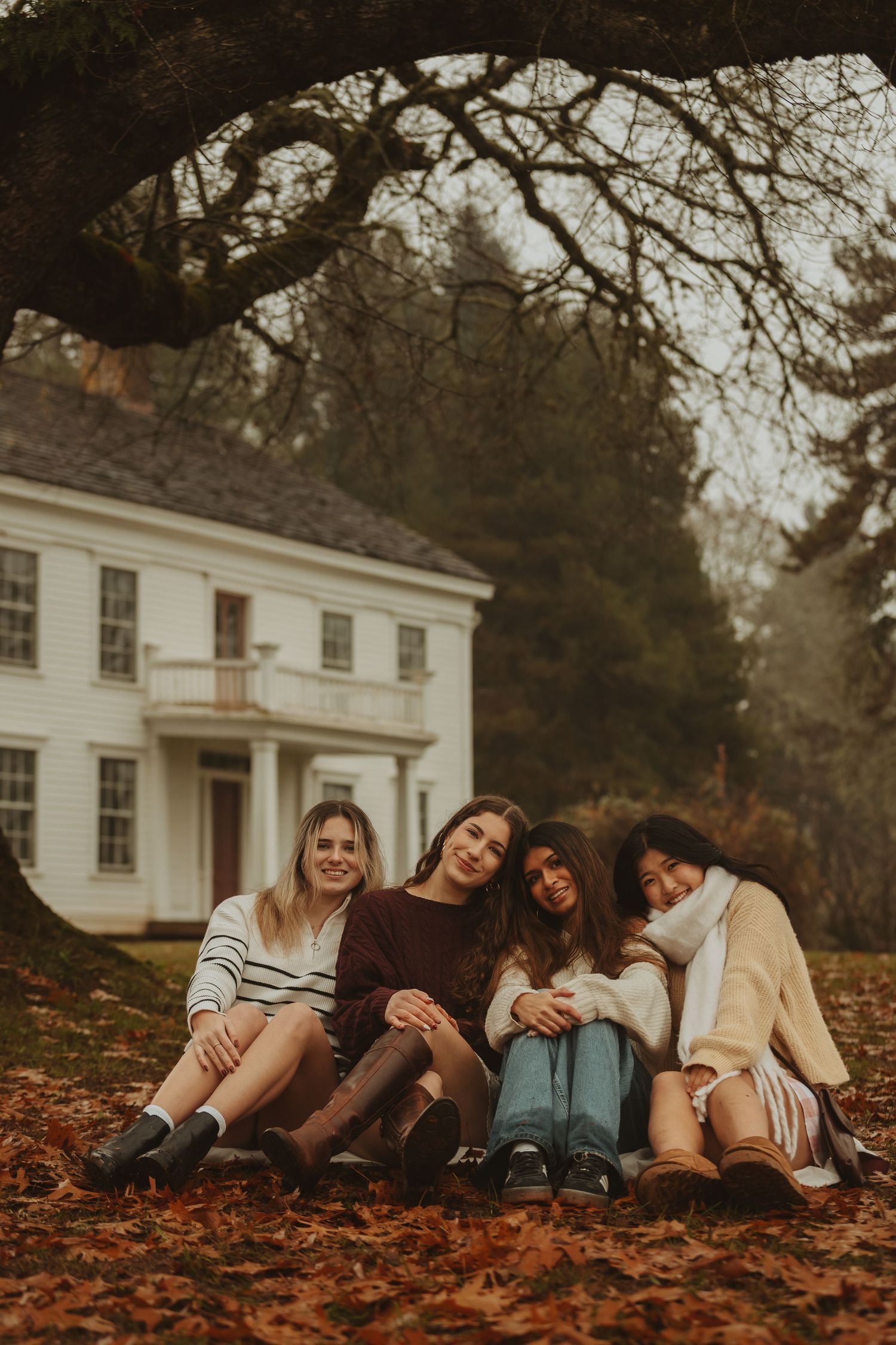 A group lies together on autumn leaves in front of a white colonial style house during a fall photo session.