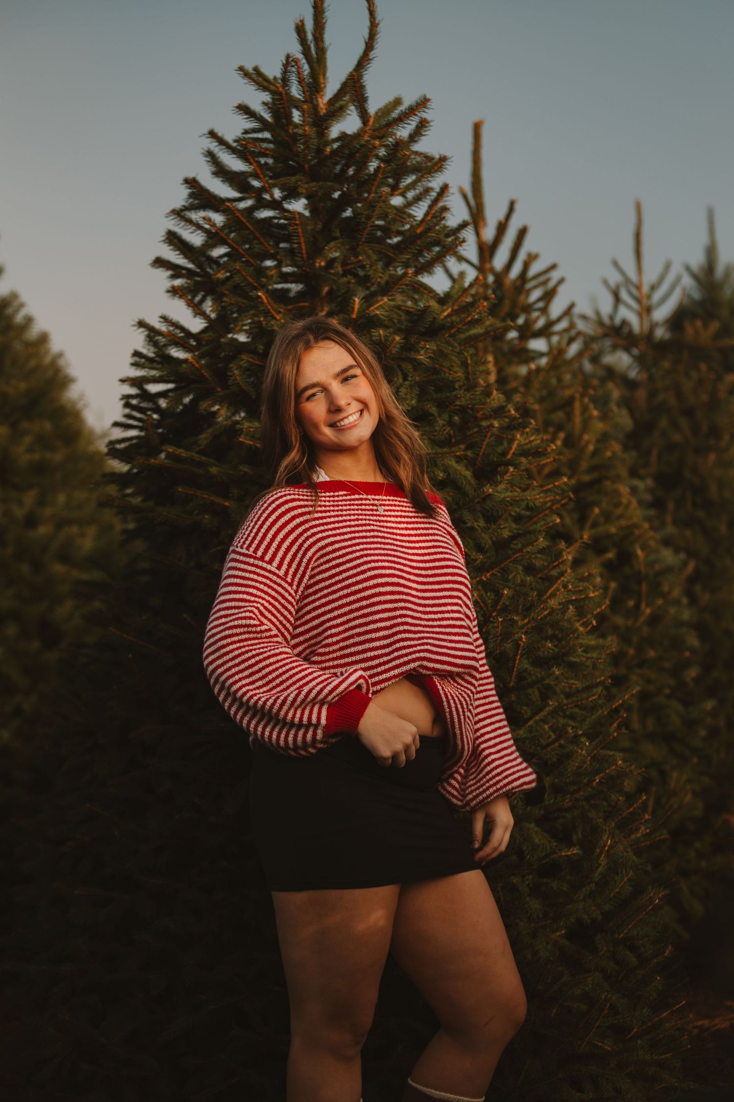 Person in striped sweater poses cheerfully against backdrop of evergreen trees.