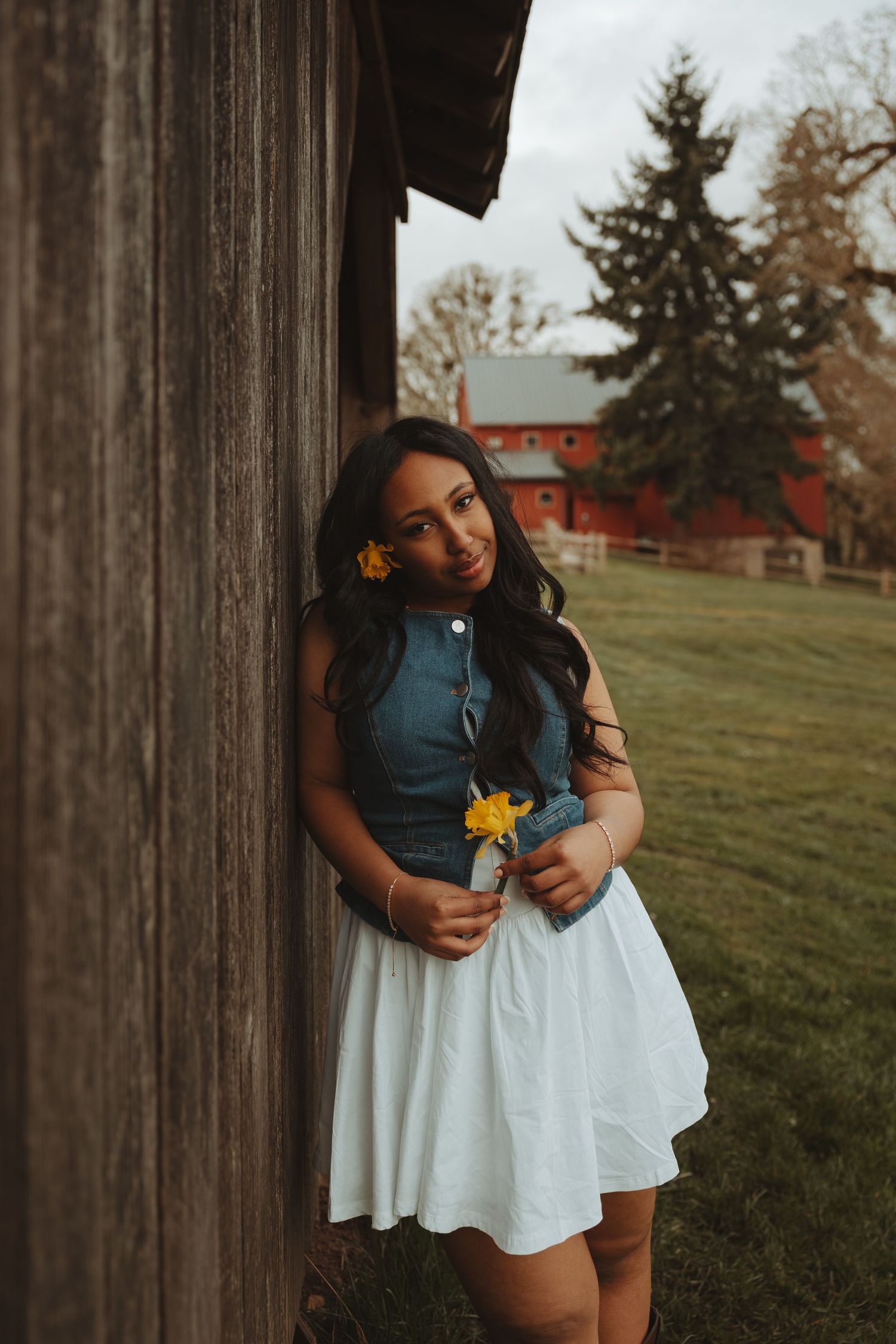 A figure in a white skirt and teal top poses against a rustic wooden post with a red barn in the background.