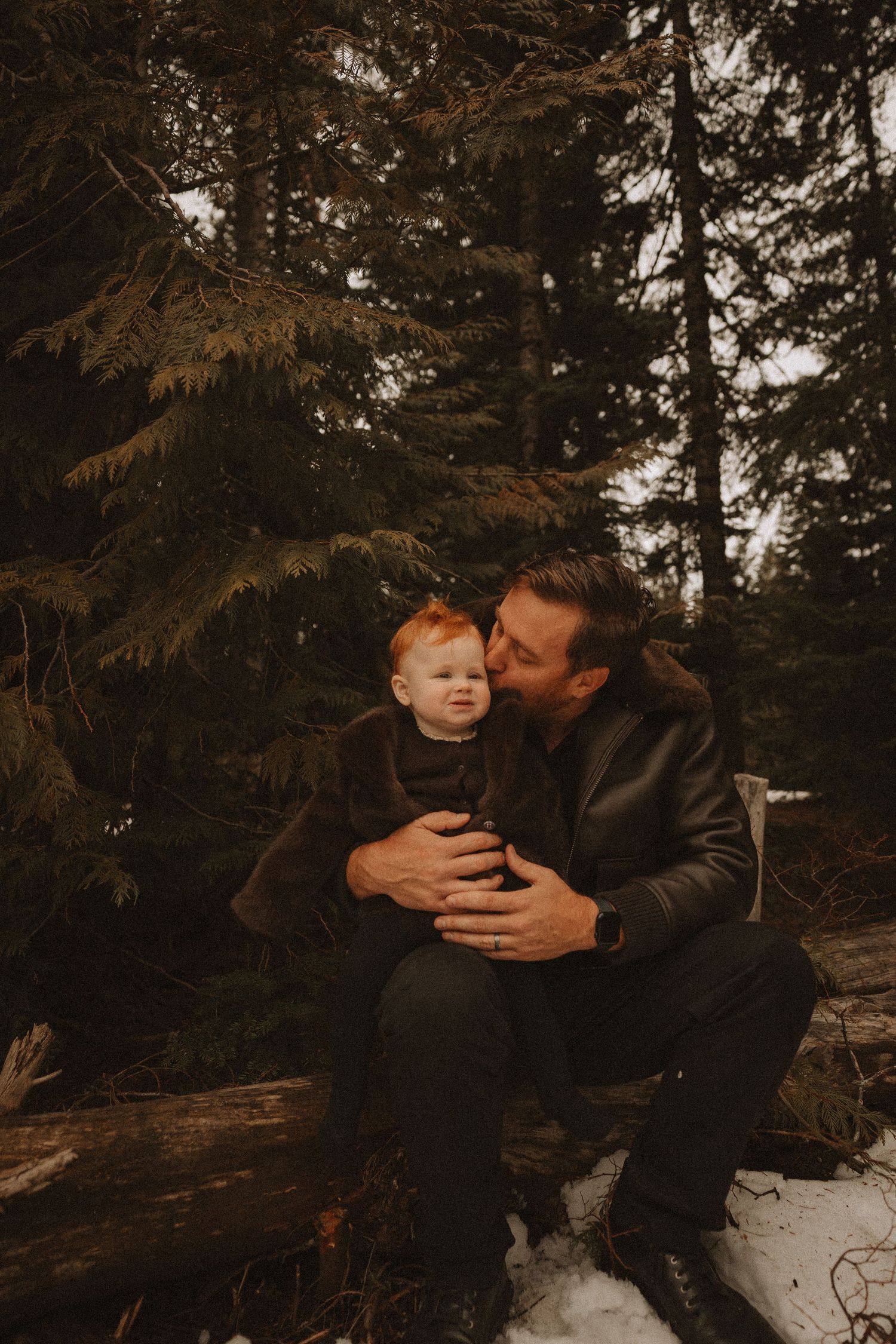 Tender moment shared between two people sitting on a fallen log in a moody forest setting at dusk.
