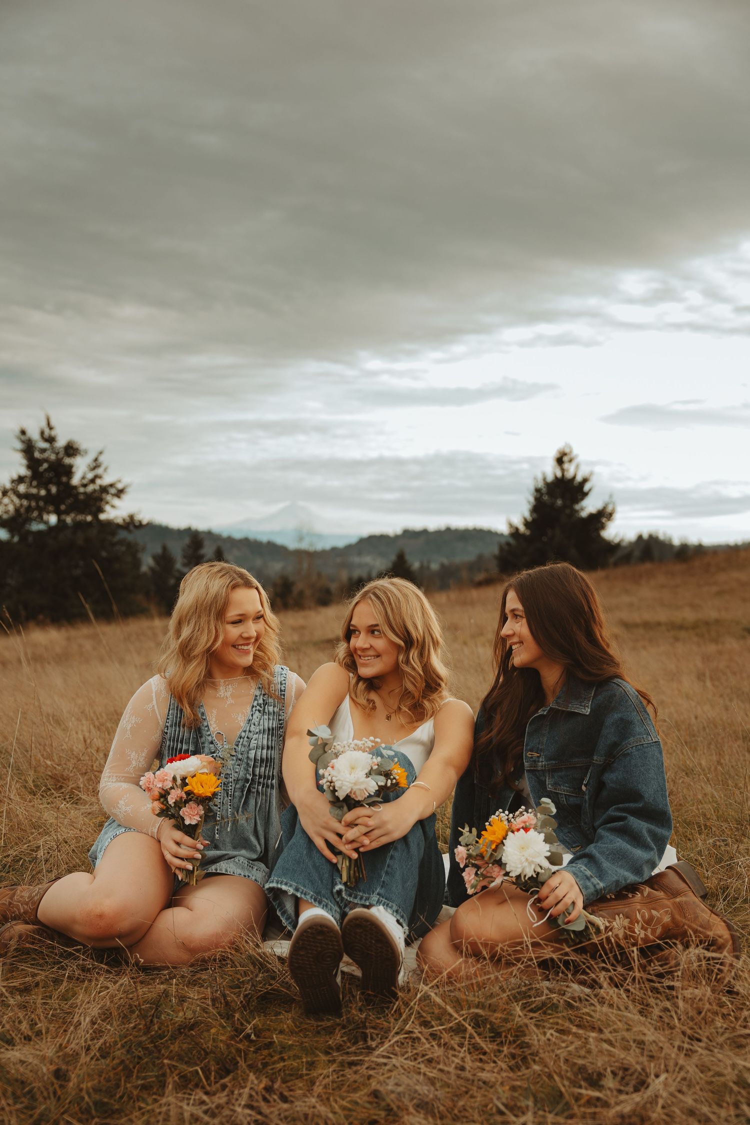 Friends lounging in a grassy field holding flowers and wearing denim and sweaters on an autumn day.