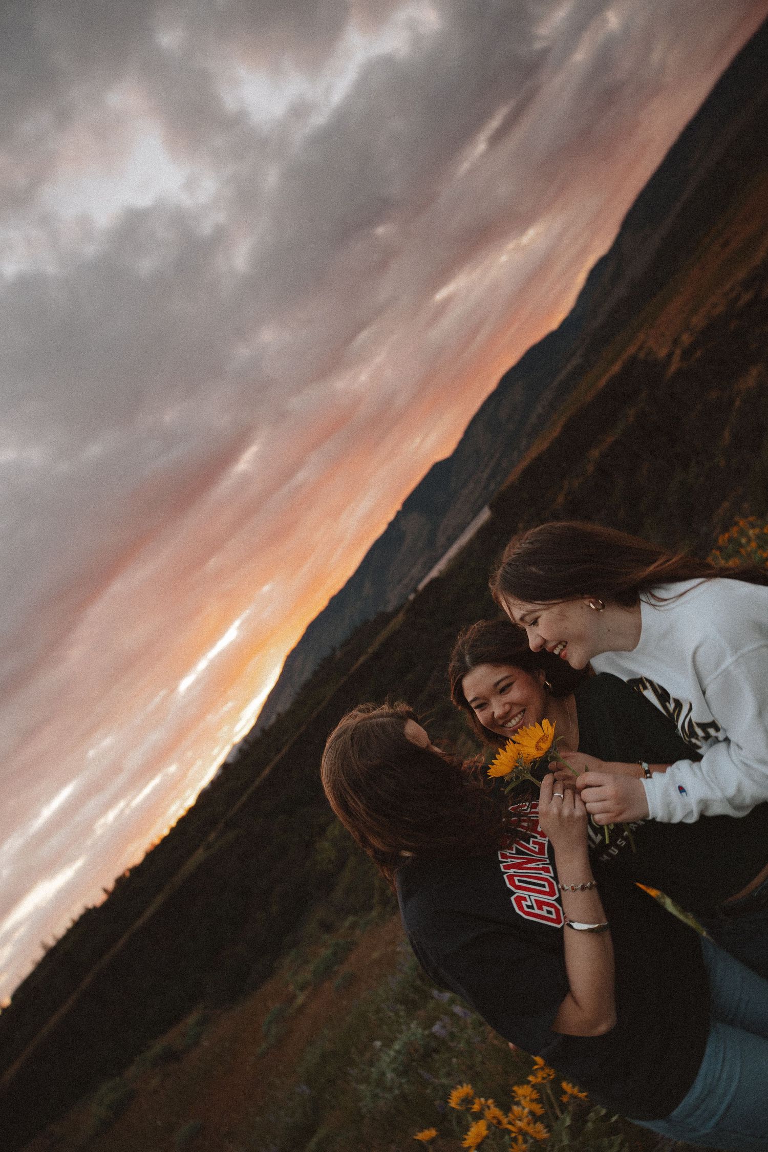 Two women sharing a tender moment holding a sunflower against a stunning colorful sunset sky over mountain landscape.