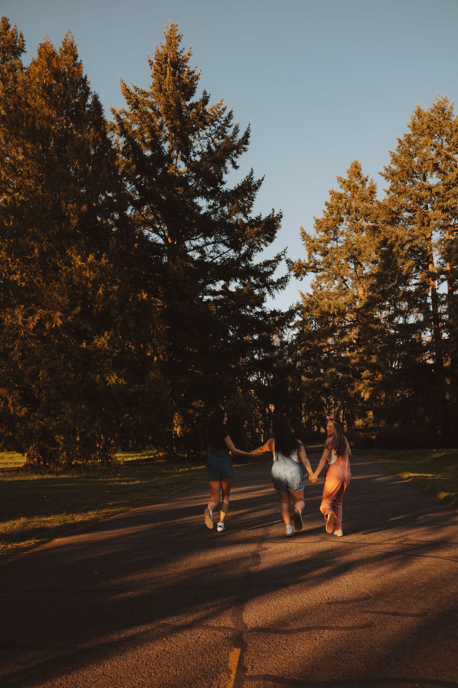 Three young women holding hands walking down a tree-lined road during golden hour sunset.