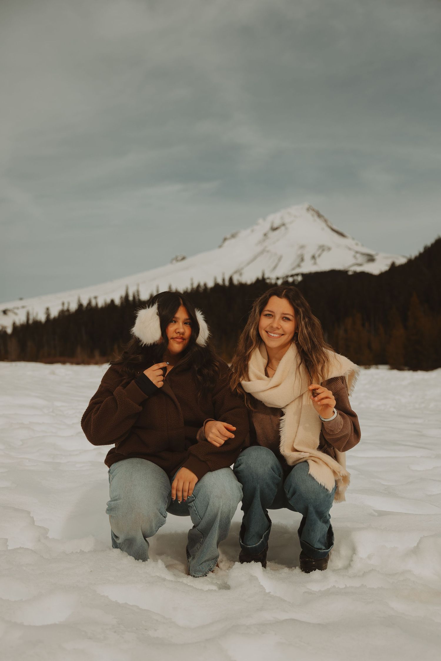 Two smiling women crouch in the snow wearing brown jackets and jeans with a mountain behind them.