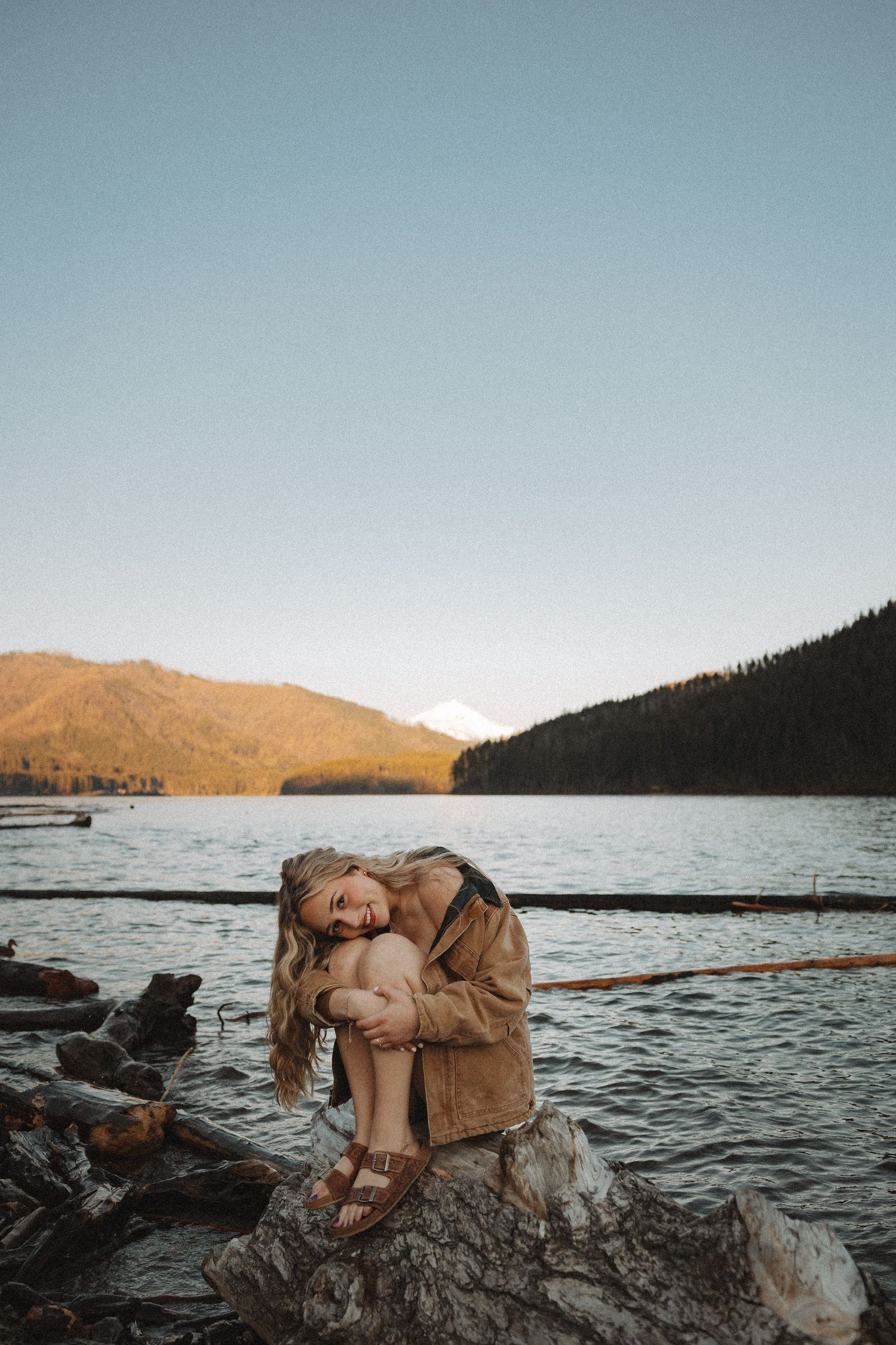 Woman in brown jacket sitting on rocks by a calm mountain lake during golden hour.