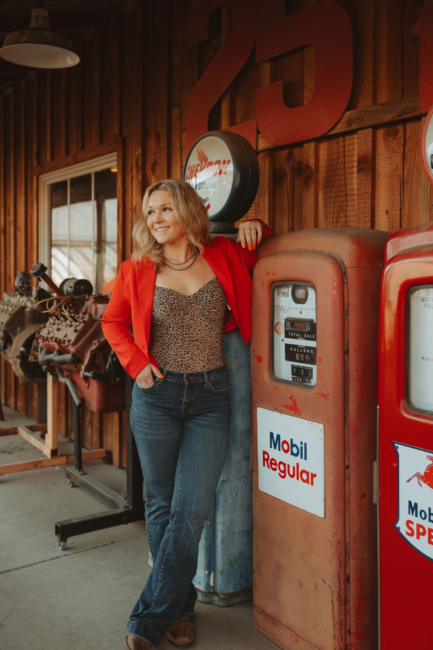 Woman in red blazer and leopard top leans against vintage Mobil Regular gas pump in rustic wooden garage.