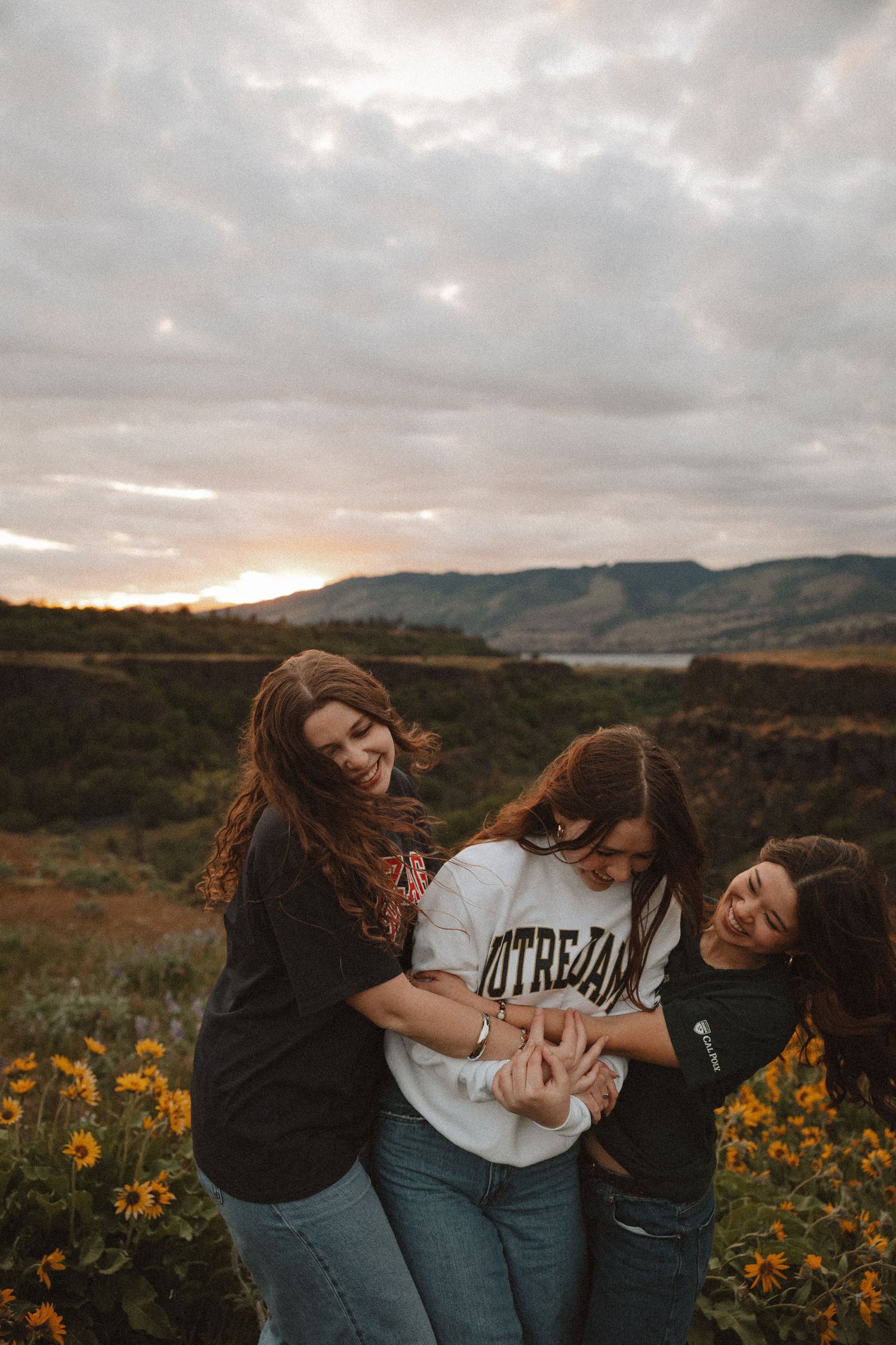 Three friends laughing and playfully embracing in a field of yellow wildflowers at sunset.