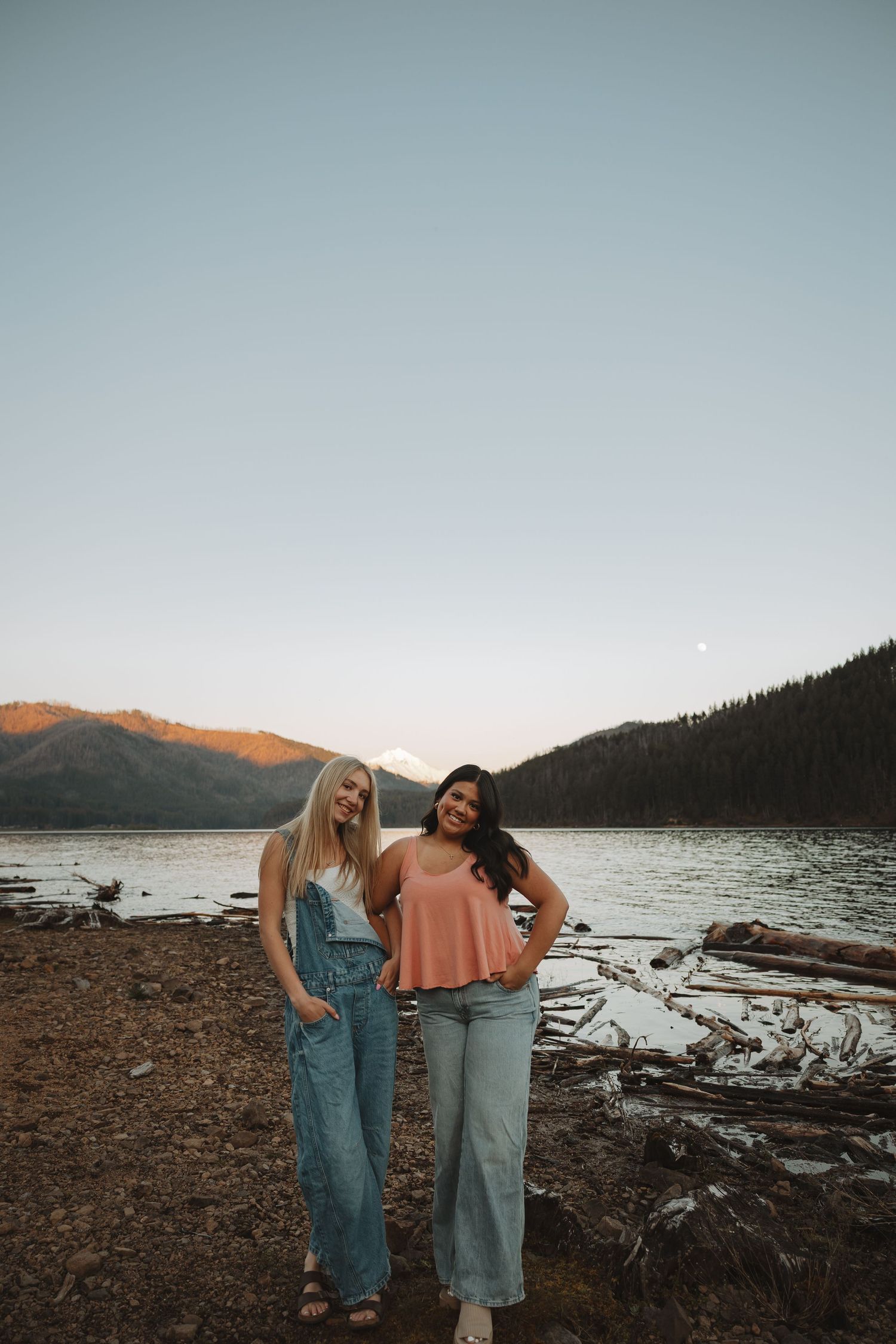 Two young women posing together on a rocky shore with mountain lake and sunset backdrop.