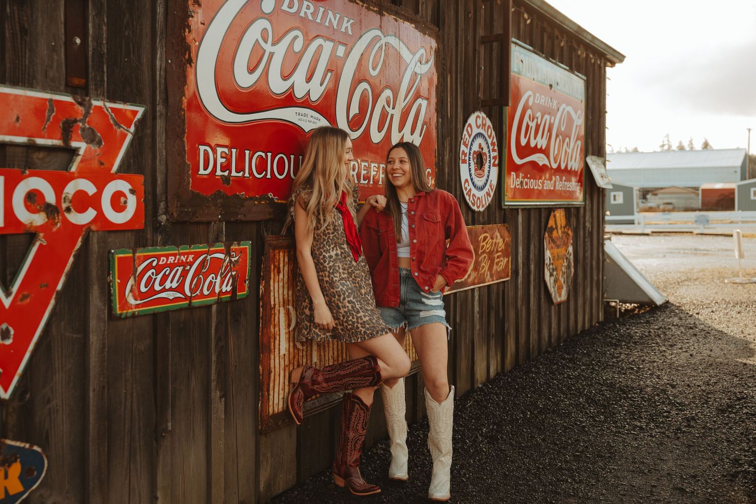 Two women pose by rustic Coca-Cola signs on a weathered barn wall, wearing Western outfits with cowboy boots.