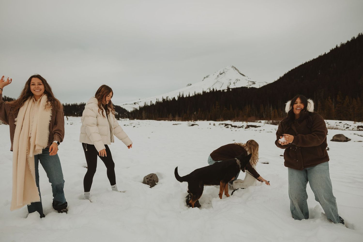 Three women playing with dogs in snowy field with mountain and pine trees in background.