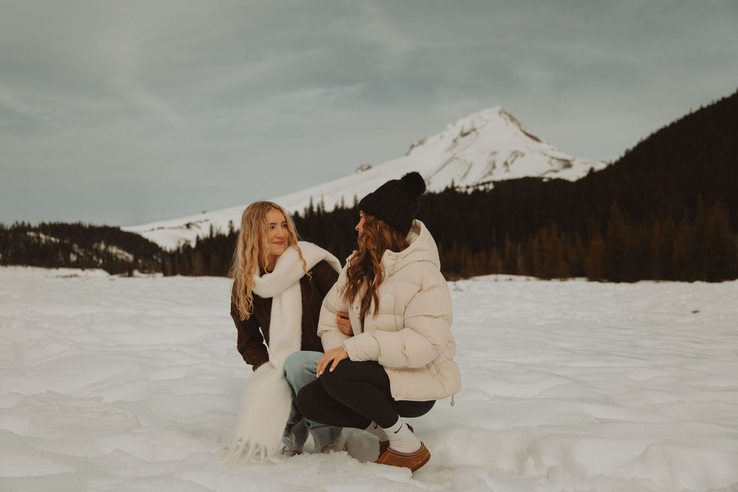 Two women sitting and kneeling in the snow, smiling at each other with a mountain backdrop.