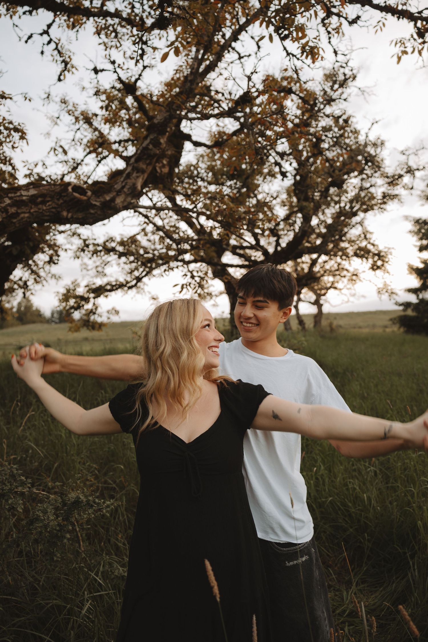 Joyful couple dancing outdoors under a large oak tree in a green meadow, woman in black dress, man in white tee.