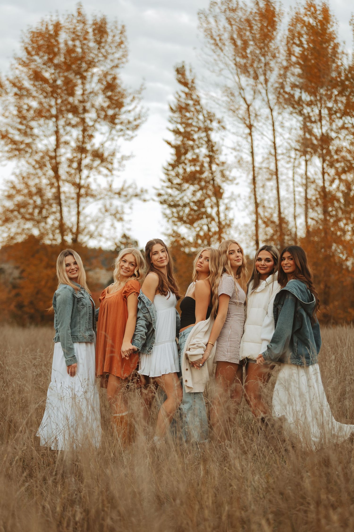 Group poses together in white dresses and fall colors against autumn trees in a field.